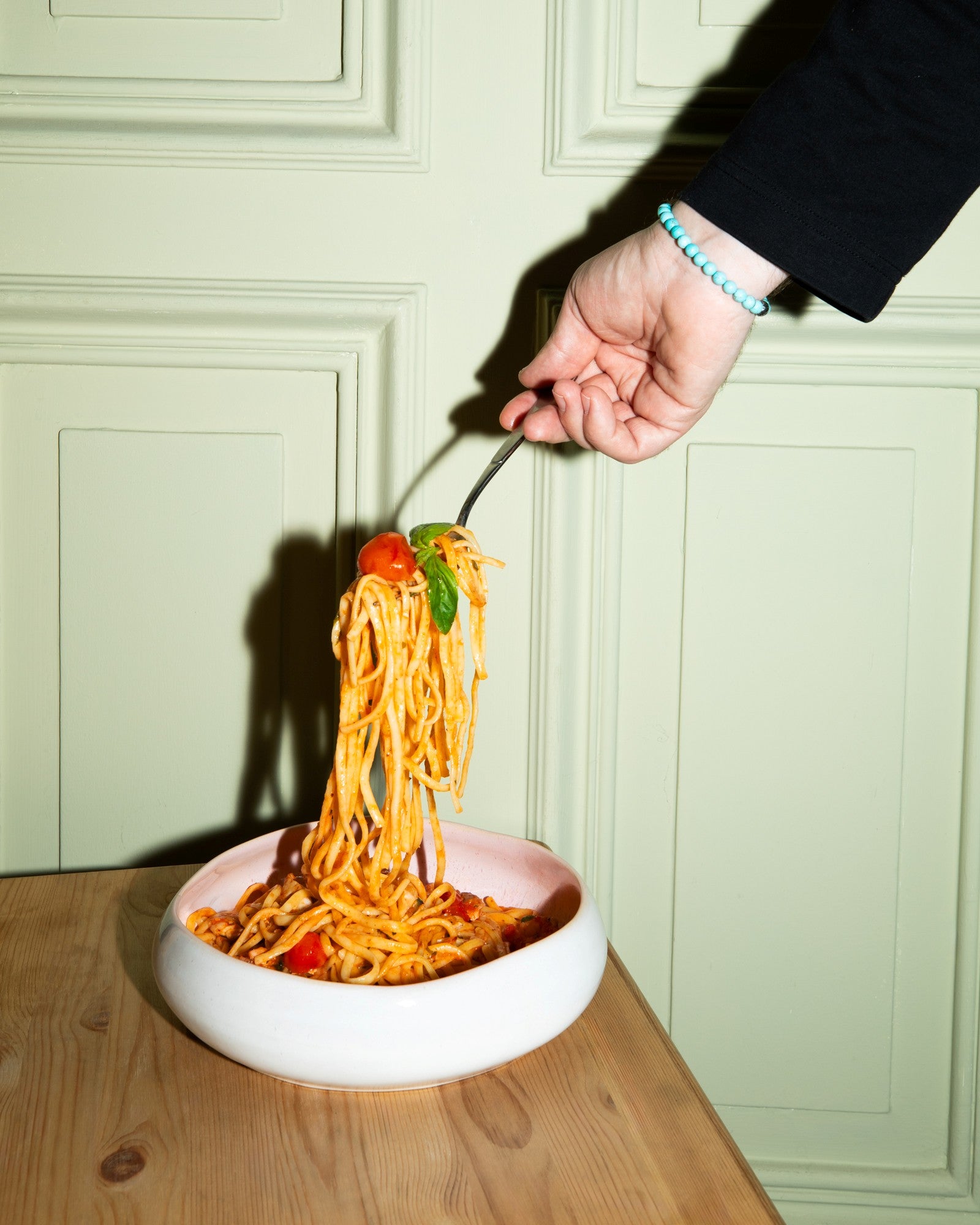 Eine Hand hält eine Gabel mit Spaghetti über einer Schüssel, vor einer pastellgrünen Wand.
