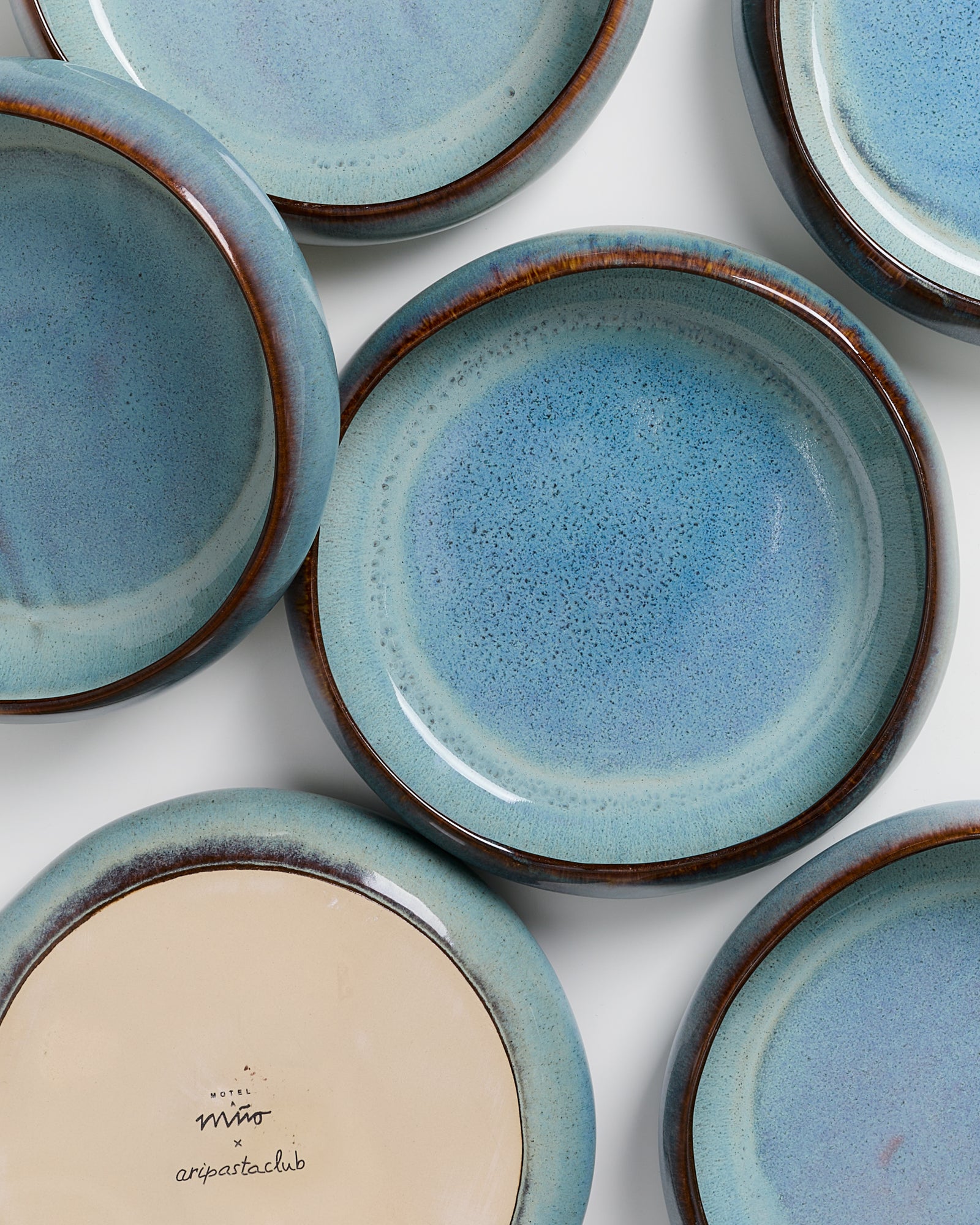 Sechs hellblaue Keramikteller mit braunem Rand, einer zeigt die Unterseite mit einer Beschriftung.