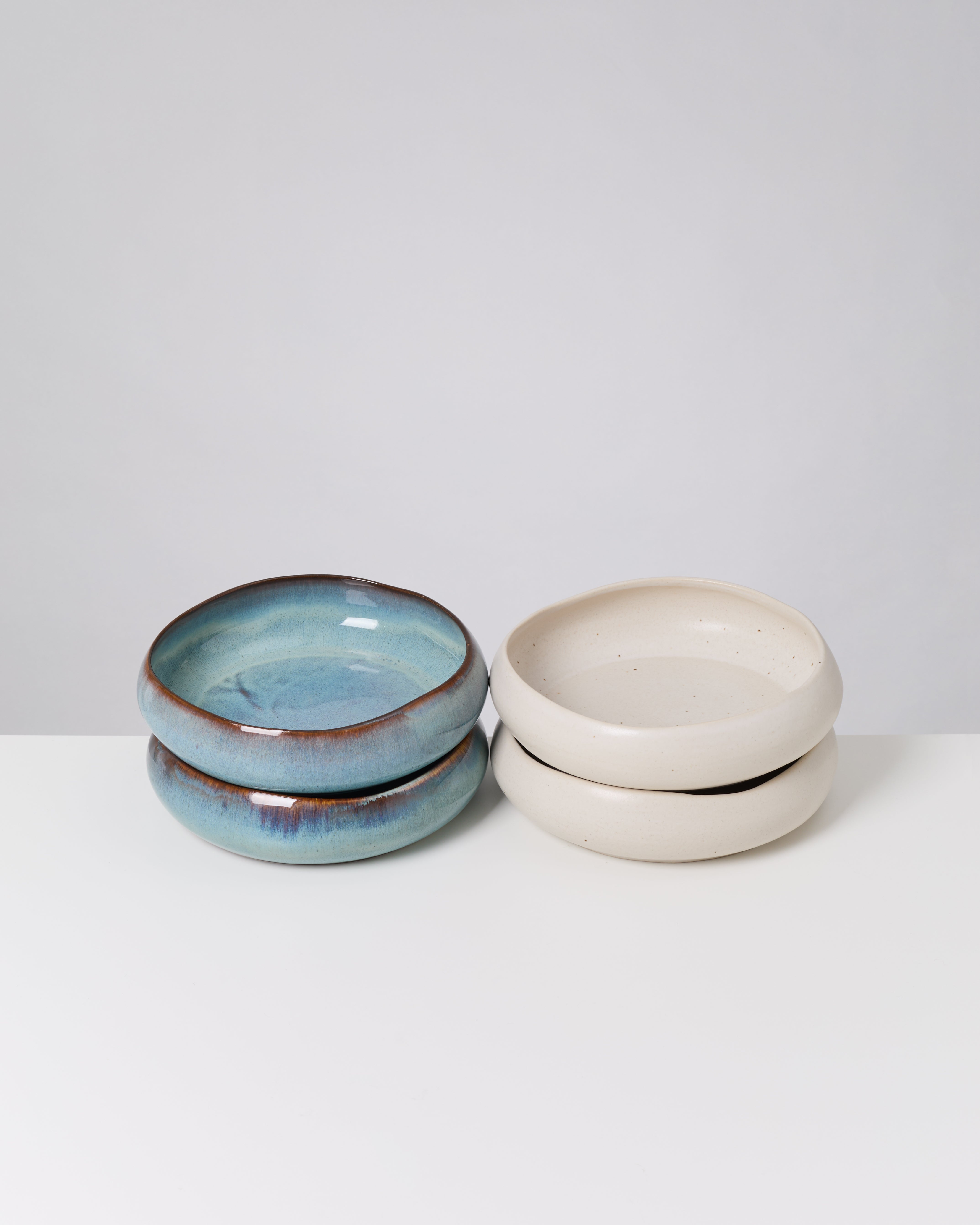 Zwei Stapel flacher, runder Keramikteller, einer in Blau, der andere in Creme, auf einem weißen Hintergrund.