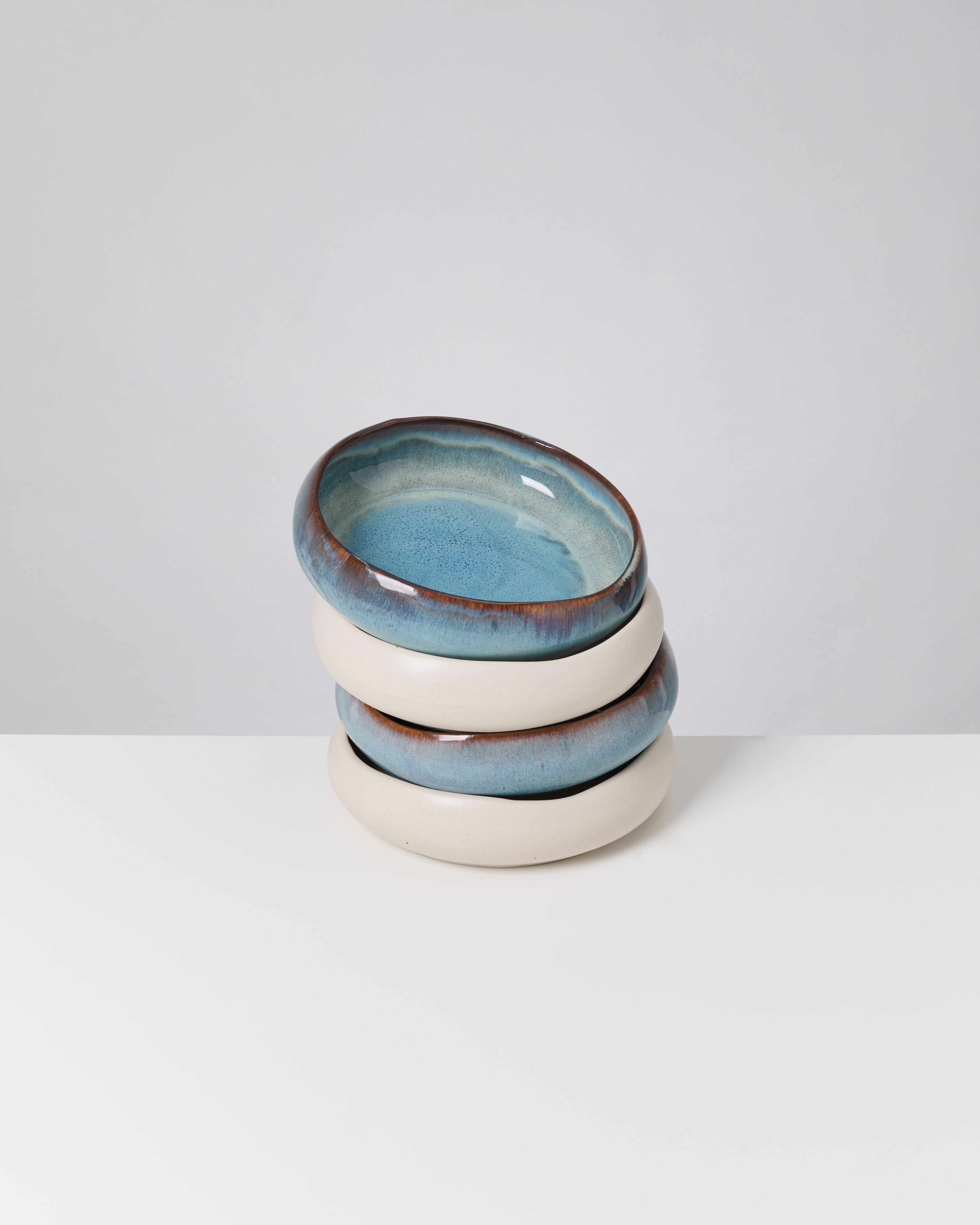 Ein Stapel von drei blauen Keramiktellern mit weißem Rand auf einem weißen Tisch vor grauem Hintergrund.