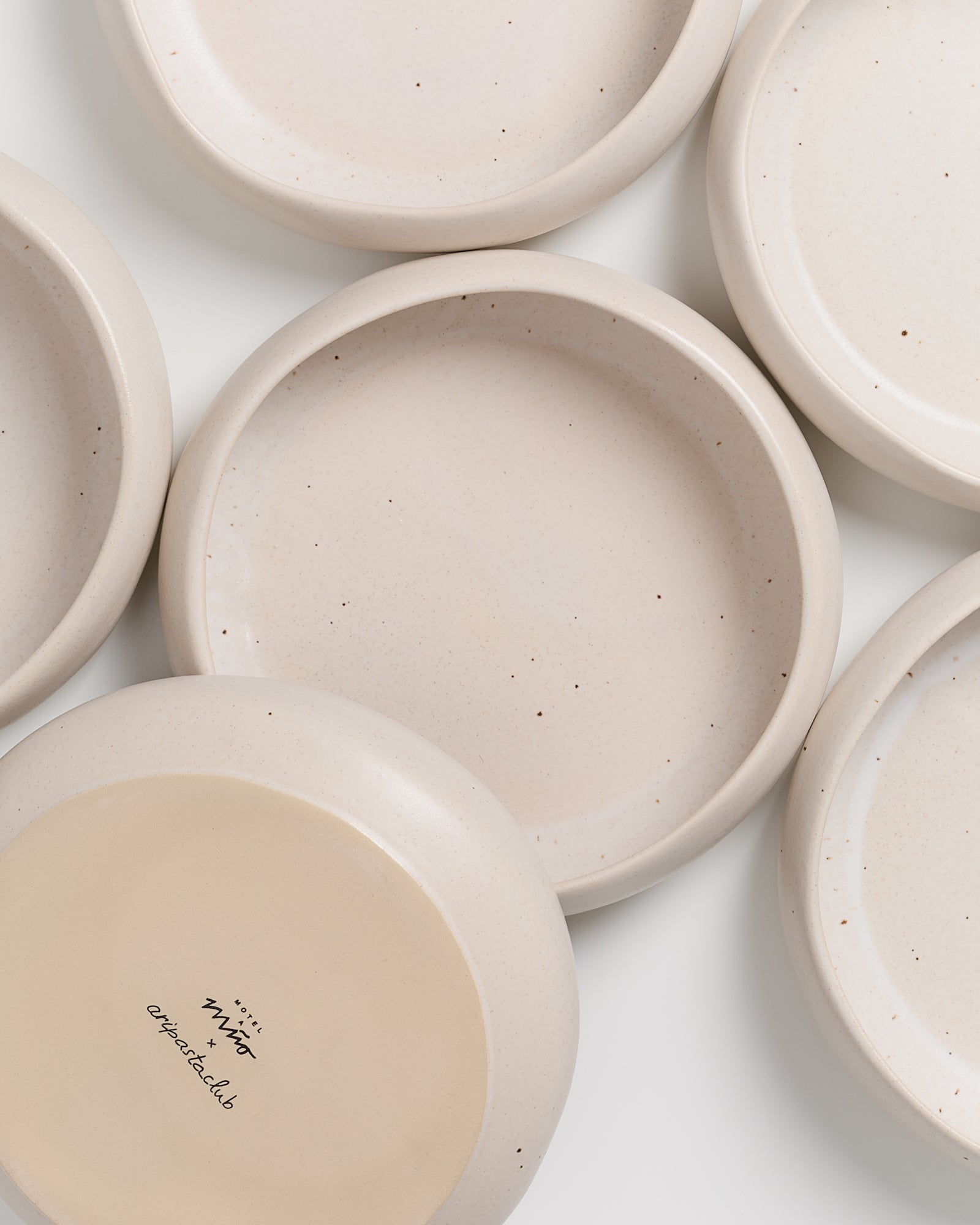 Sechs beige, runde Keramikteller mit feinen Sprenkeln, einer zeigt die Unterseite mit einer Aufschrift.