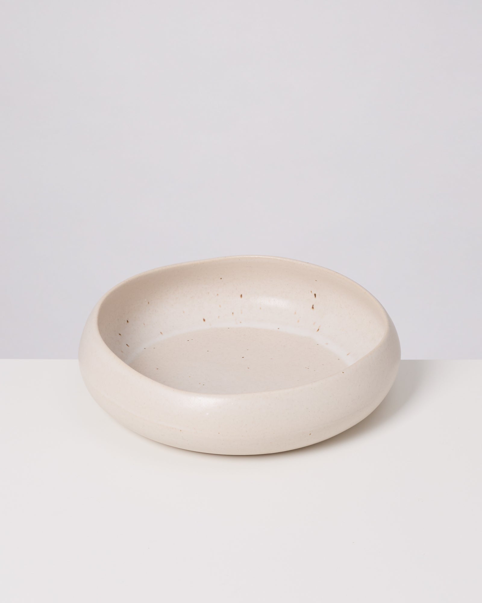 Cremefarbene, flache Keramikschüssel mit matter Oberfläche und kleinen Sprenkeln auf einem weißen Hintergrund.