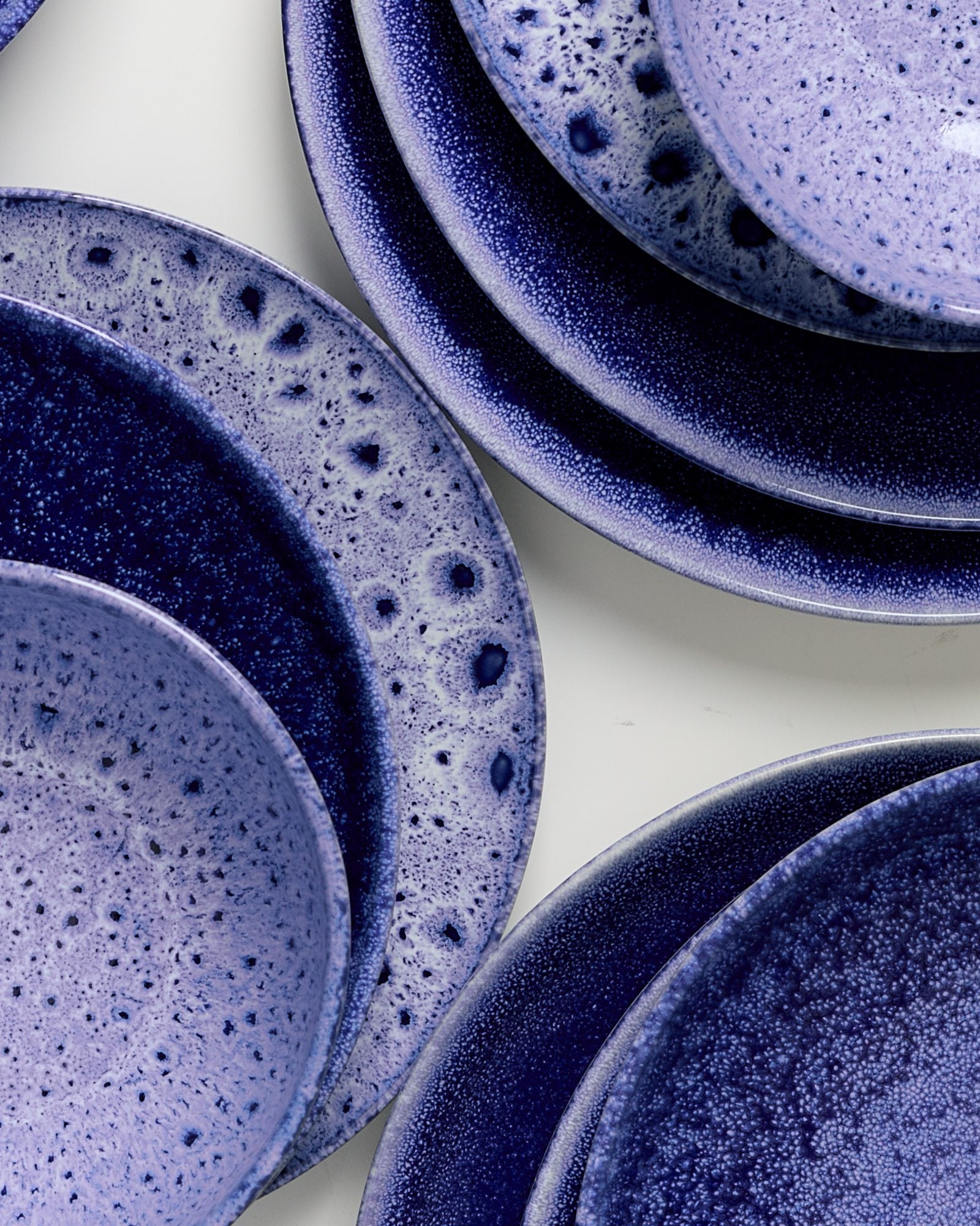 Violett-blaue, gesprenkelte Keramikteller in verschiedenen Größen überlappen sich auf einer weißen Oberfläche.