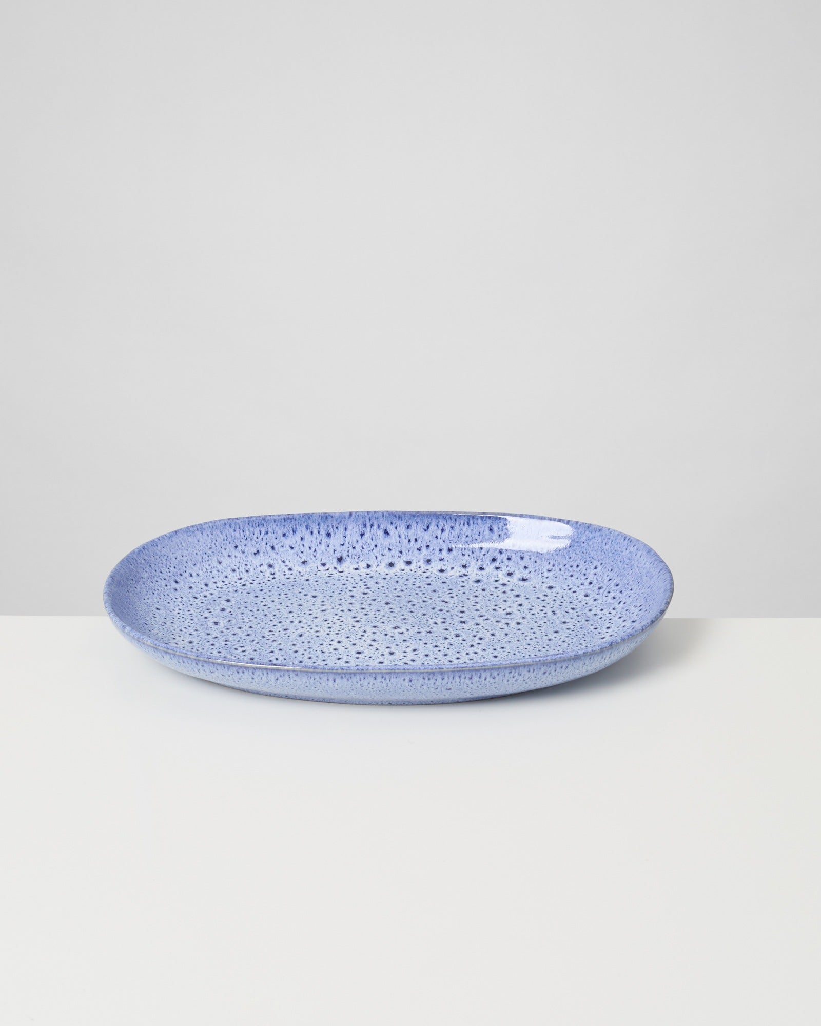 Blauer, ovaler Teller mit gesprenkeltem Muster auf einem weißen Tisch, minimalistisch vor grauem Hintergrund dargestellt.