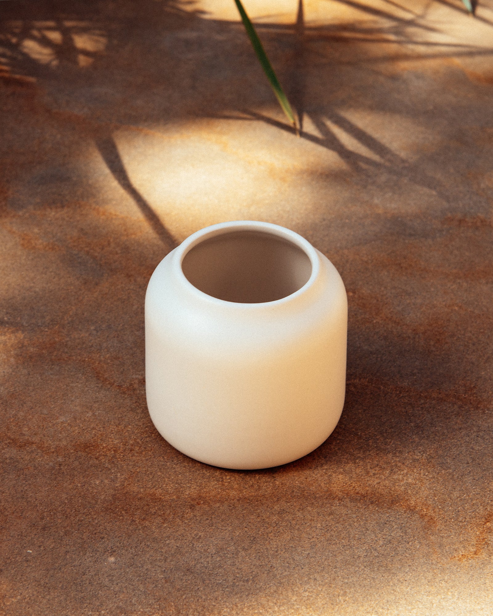 Beiger Keramiktopf auf sandigem Untergrund mit Schatten von Pflanzen.