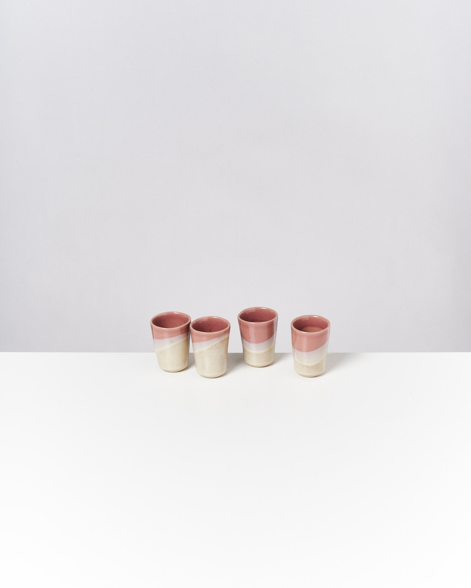 Vier zweifarbige, beige und rosa, Keramikbecher stehen auf einer weißen Oberfläche vor grauem Hintergrund.