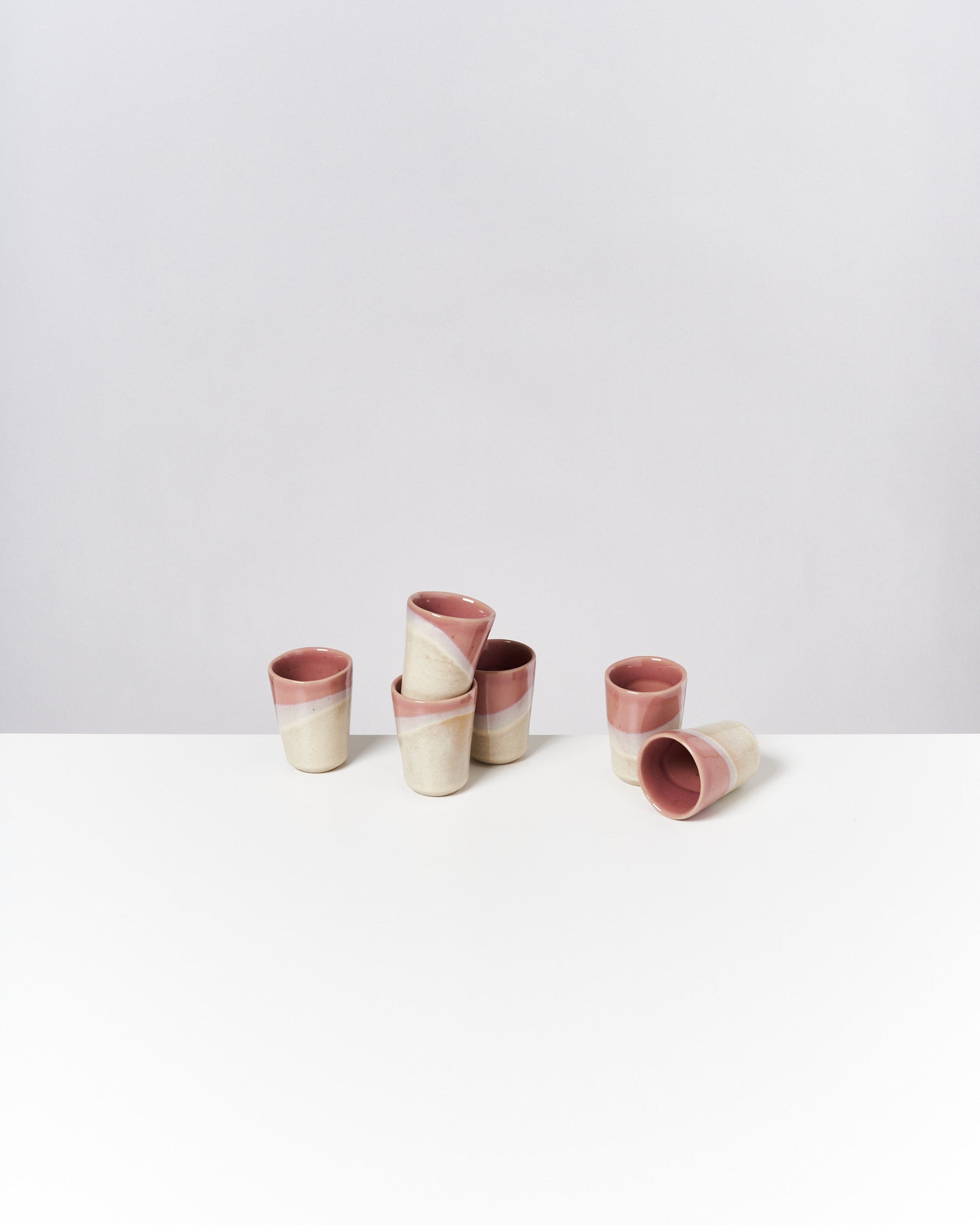 Sechs rosa-weiße Keramikbecher stehen und liegen auf einem weißen Tisch vor einem grauen Hintergrund.