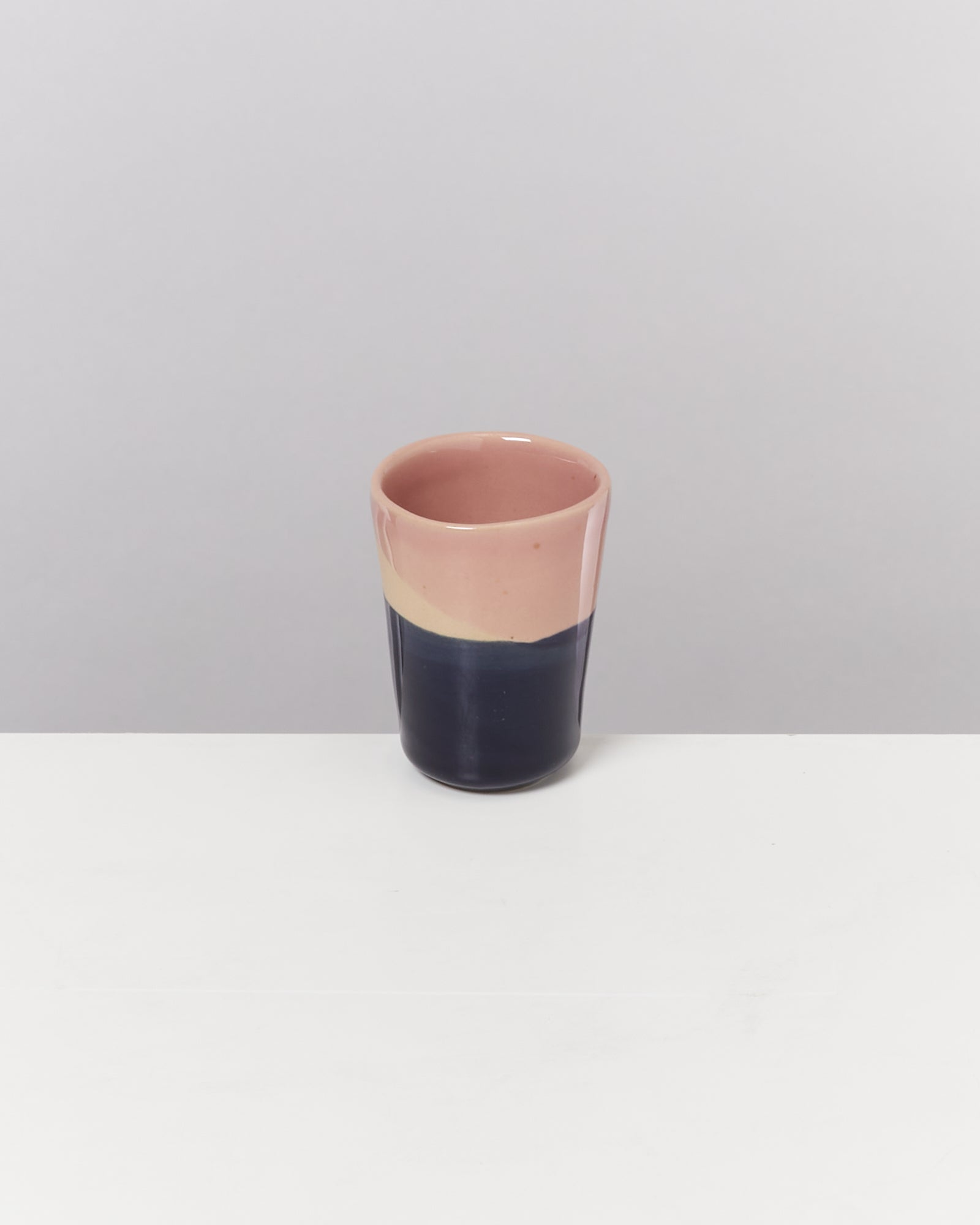 Ein zweifarbiger Keramikbecher, oben rosa und unten dunkelblau, steht auf einem weißen Tisch vor grauem Hintergrund.