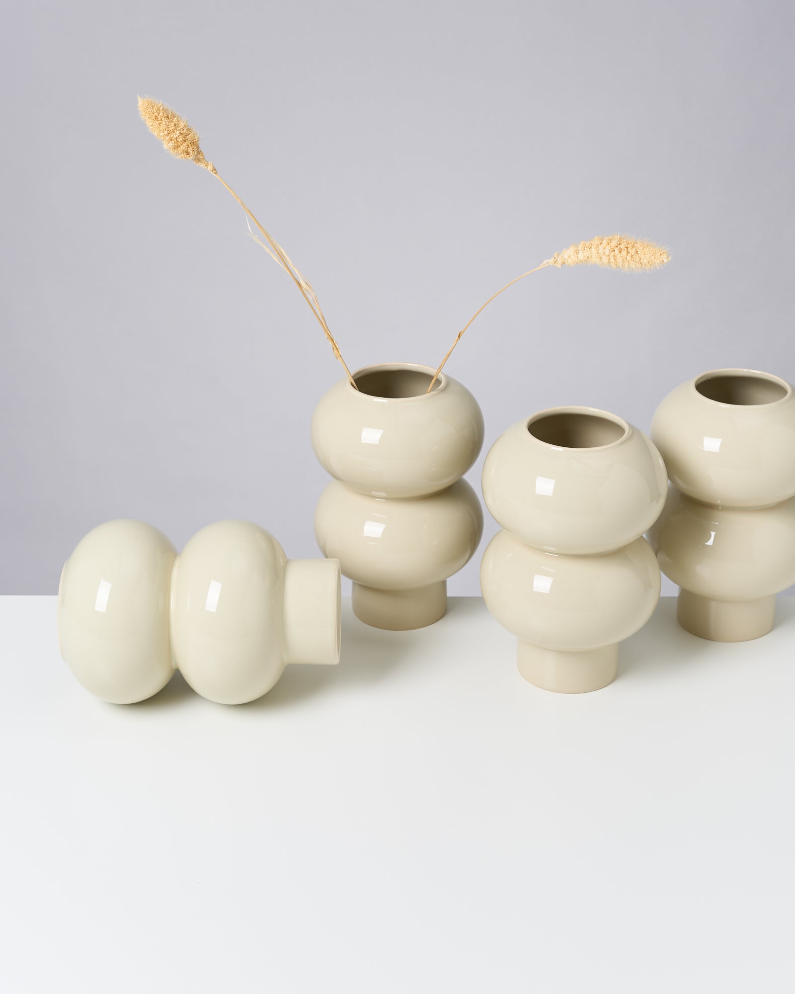 Beige, bauchige Vasen auf weißem Tisch; eine Vase liegt, zwei haben getrocknete Gräser.