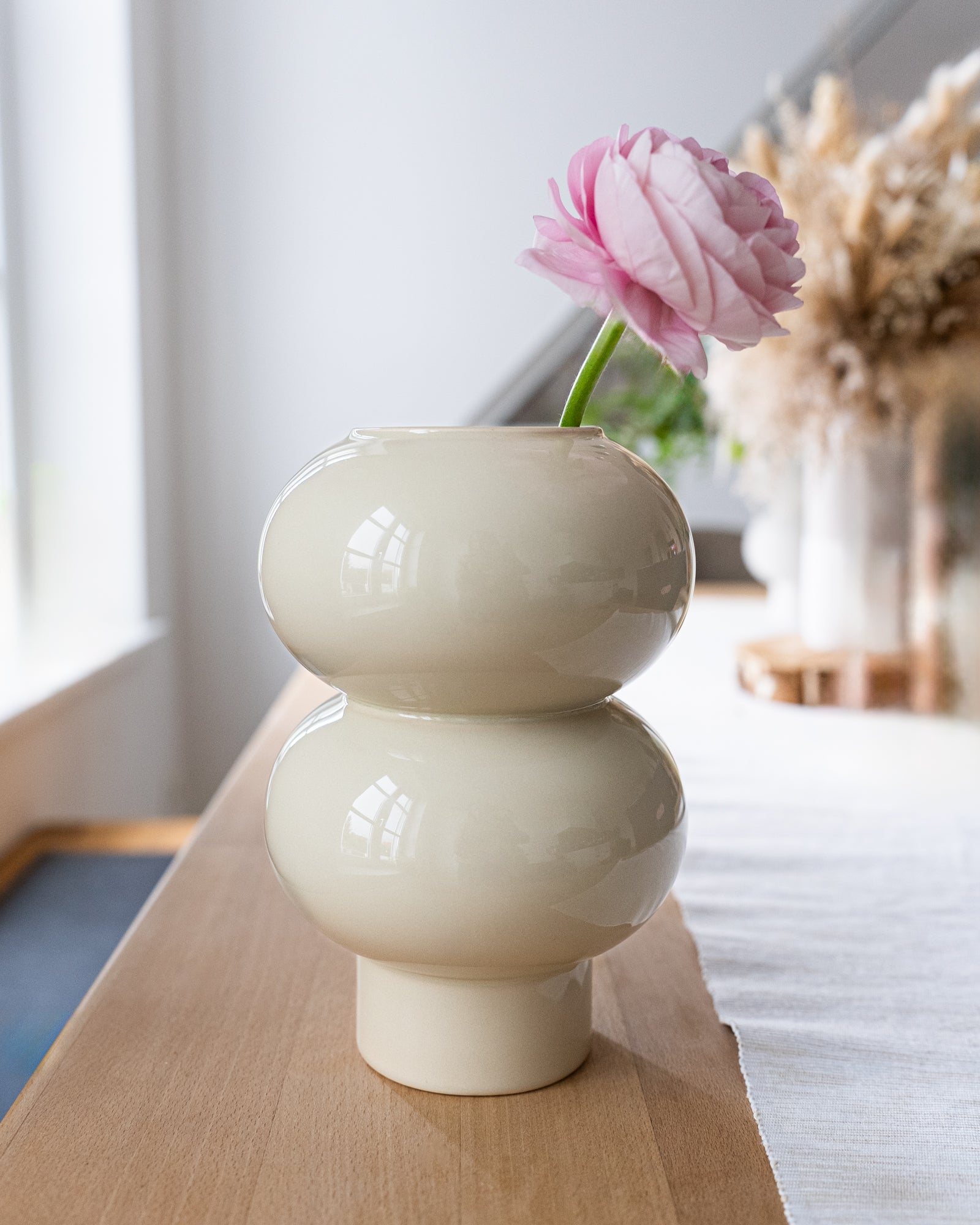 Zweistufige cremefarbene Vase mit rosafarbener Blume auf Holztisch, im Hintergrund trockene Gräser.