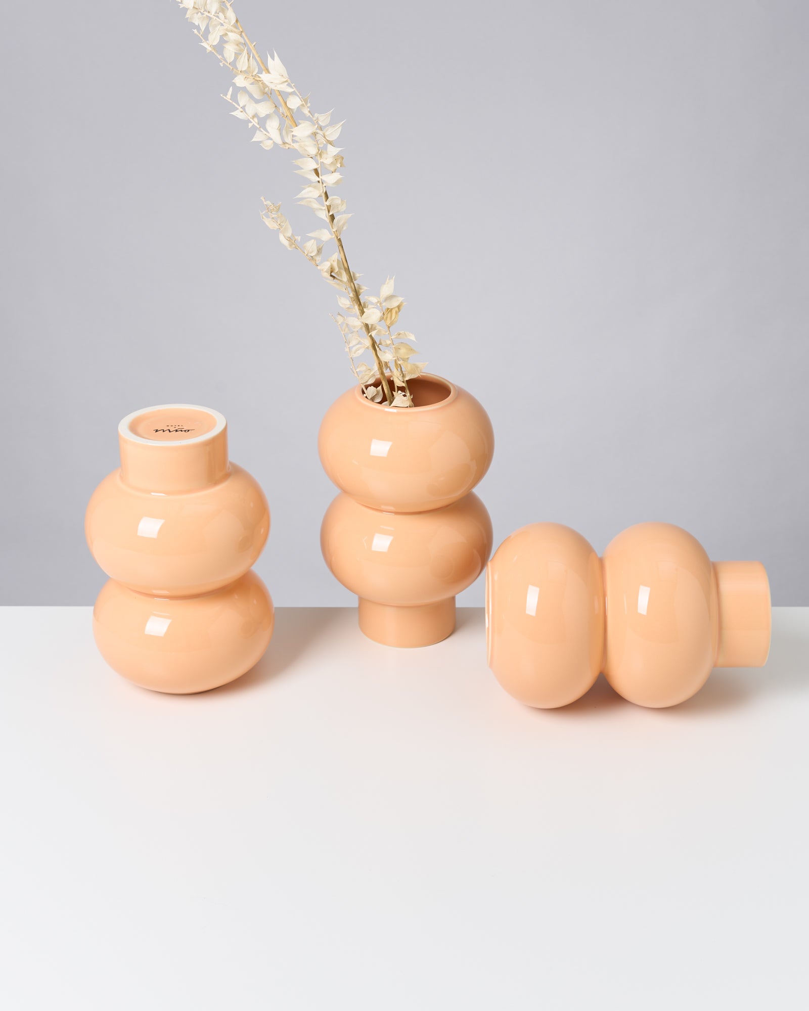 Drei beige, kugelförmige Vasen auf weißem Tisch, eine mit getrockneten Blumen gefüllt.