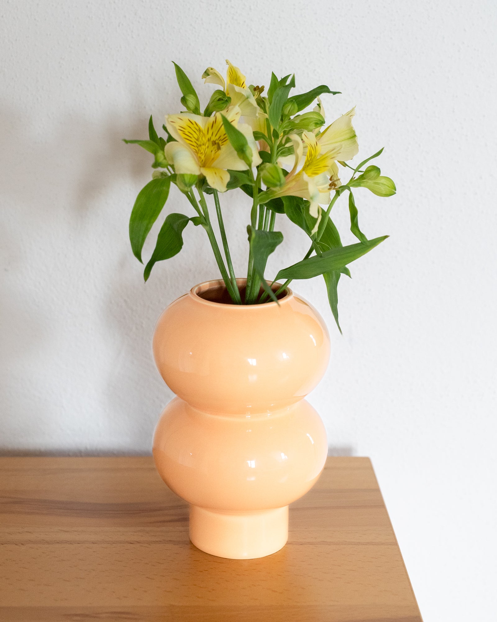 Pfirsichfarbene Vase mit gelben Lilien und grünen Blättern steht auf einem Holztisch vor einer weißen Wand.