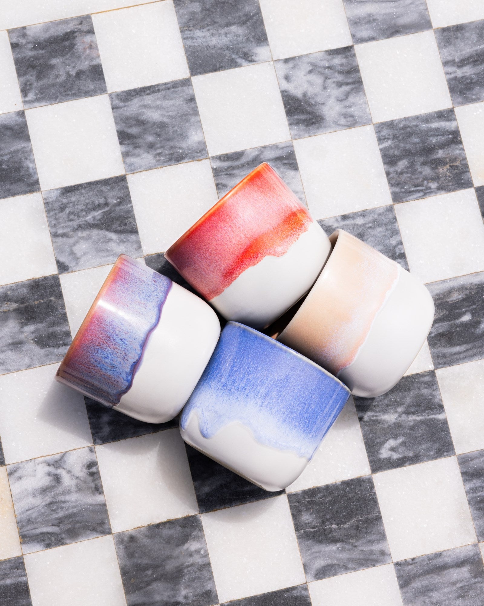 Vier bunte Keramikbecher liegen auf einem schwarz-weiß karierten Marmorboden.