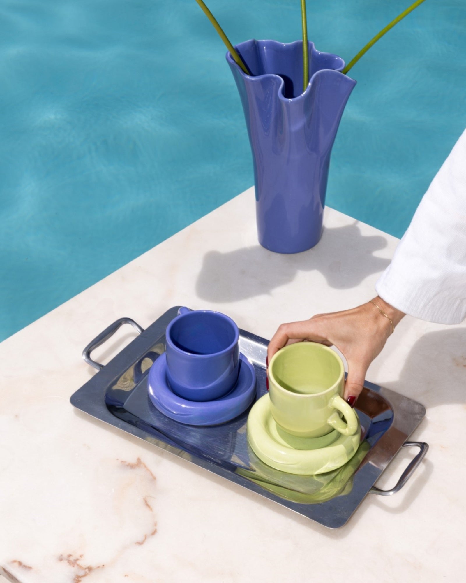 Eine Person stellt bunte Tassen auf einem silbernen Tablett neben einem Pool und einer blauen Vase mit Blumen ab.