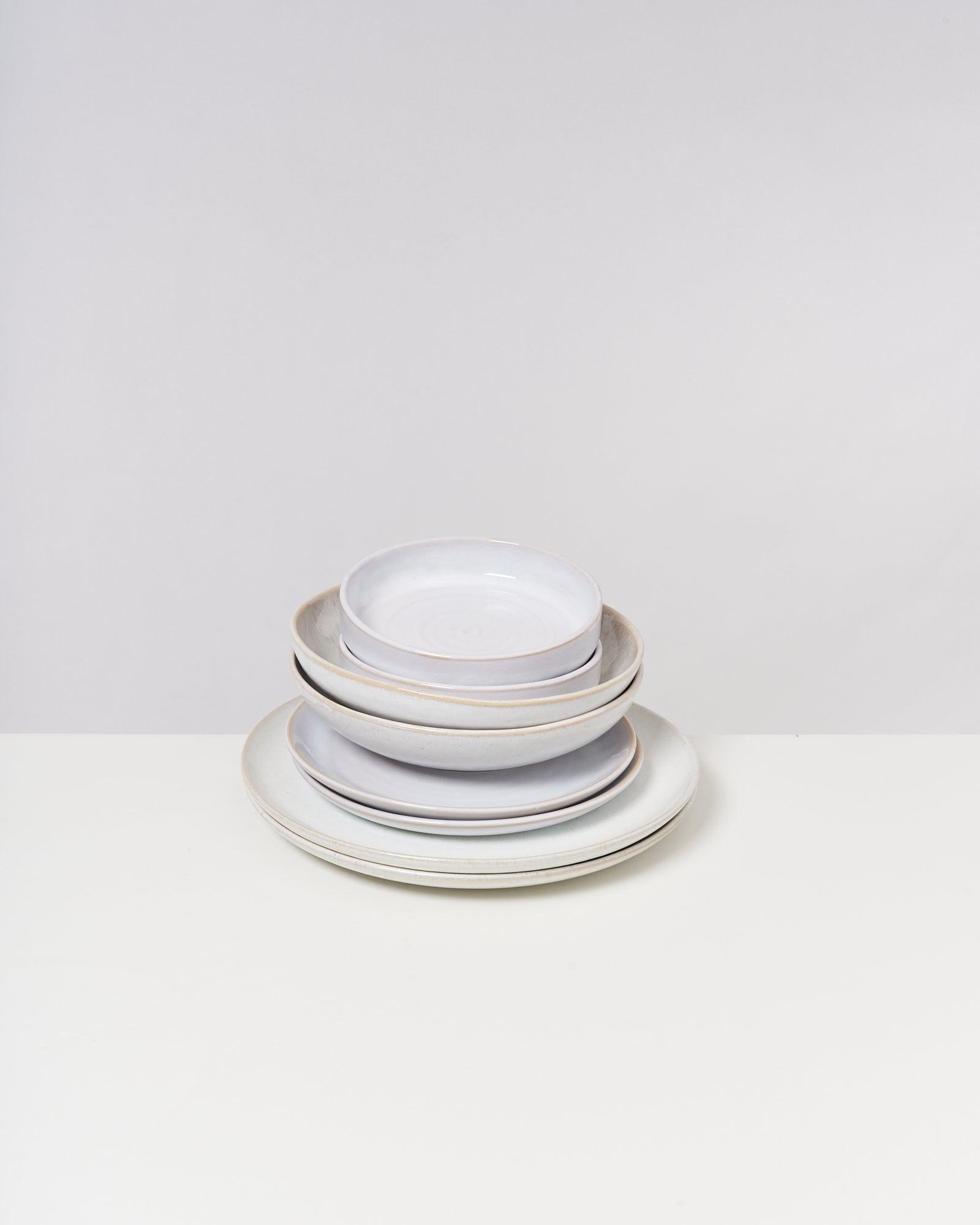 Ein Stapel weißer Keramikteller und -schüsseln auf einem weißen Tisch vor einem schlichten Hintergrund.