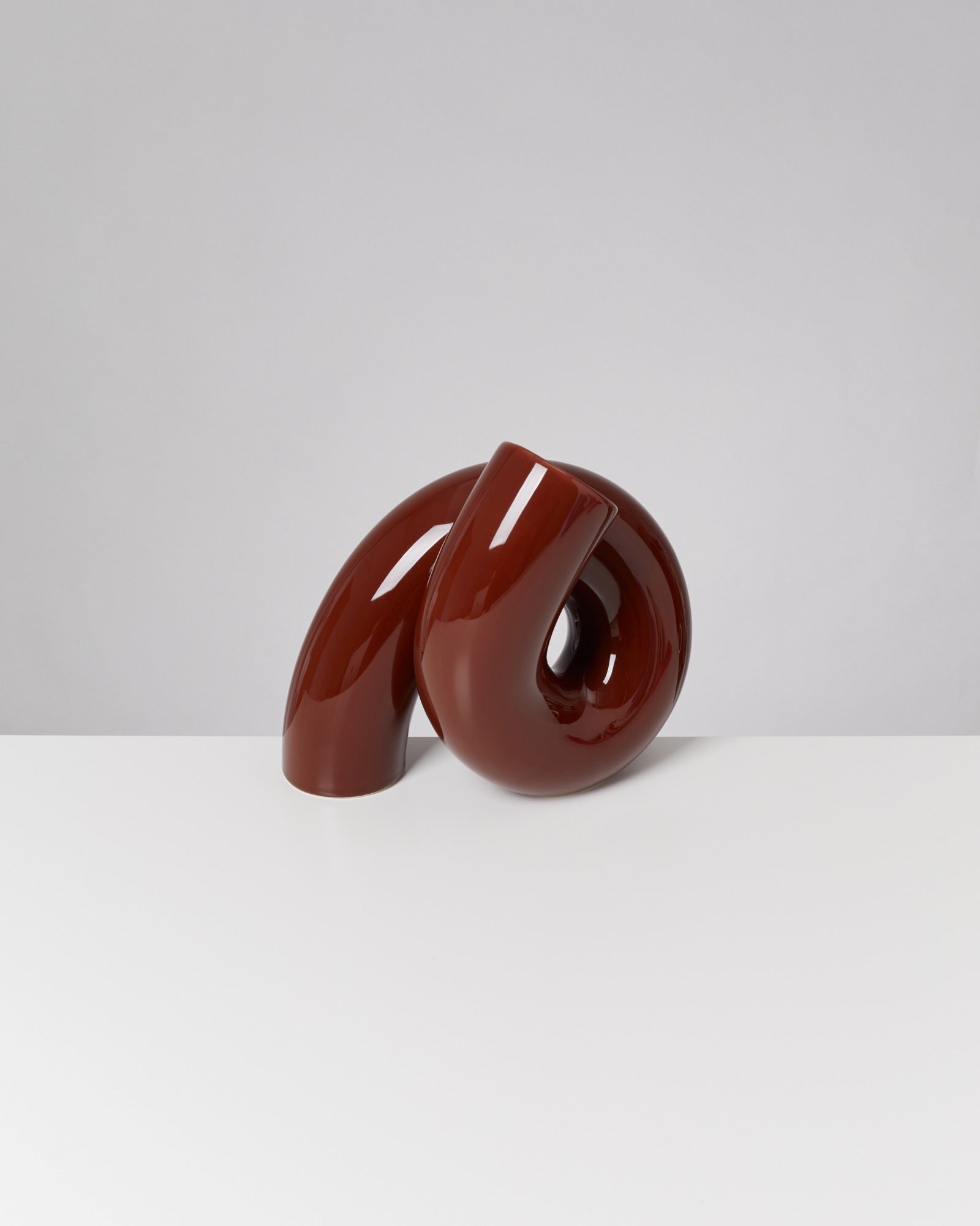 Braune, glänzende, abstrakte Keramikskulptur steht auf einem weißen Tisch vor einem grauen Hintergrund.