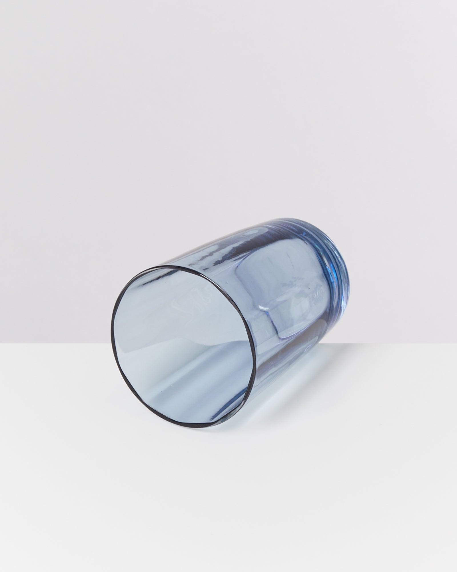 Ein blauer, liegender Glasbecher auf einer weißen Oberfläche vor neutralem Hintergrund.