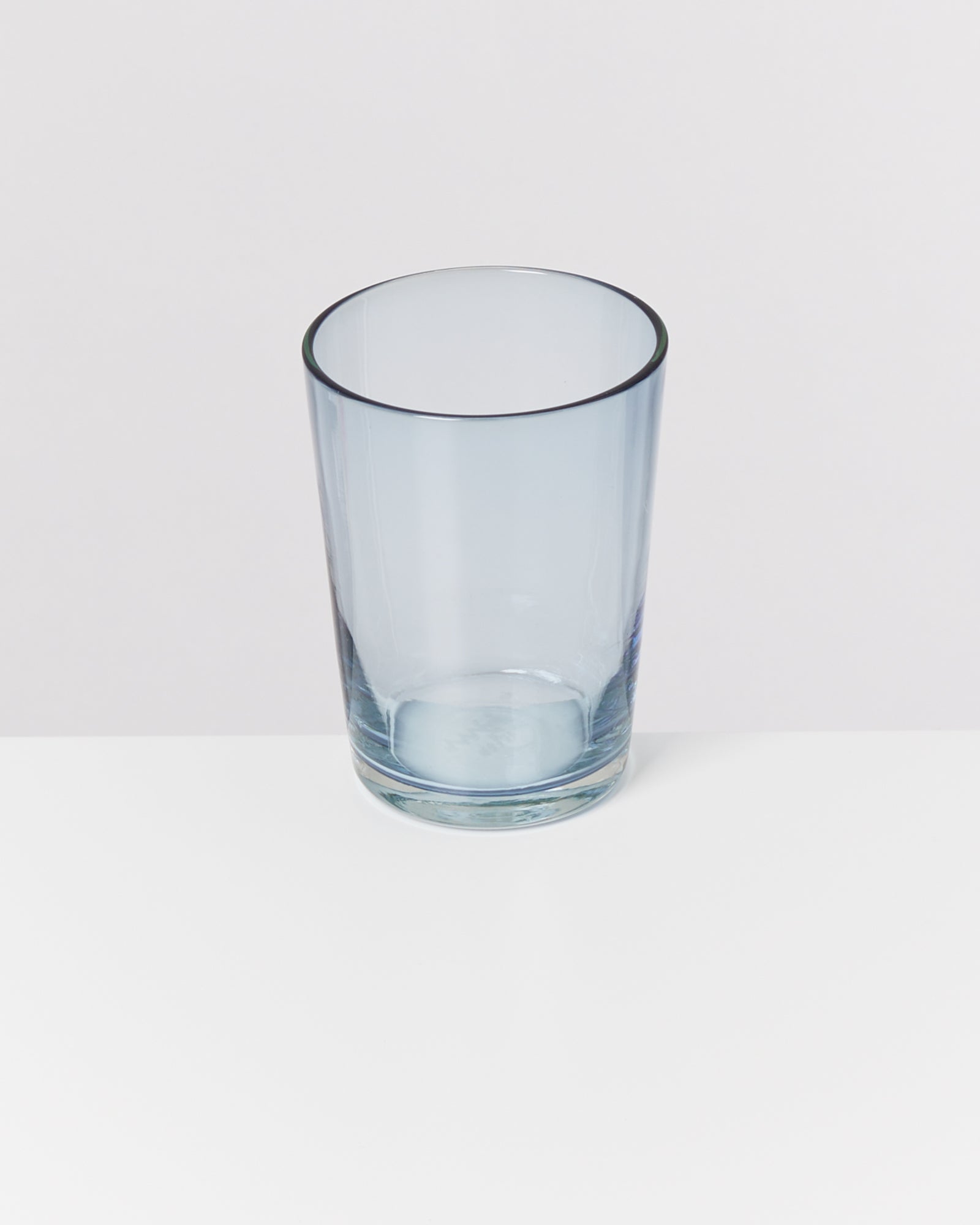 Ein leeres, durchsichtiges Glas steht auf einem weißen Untergrund vor einem hellgrauen Hintergrund.