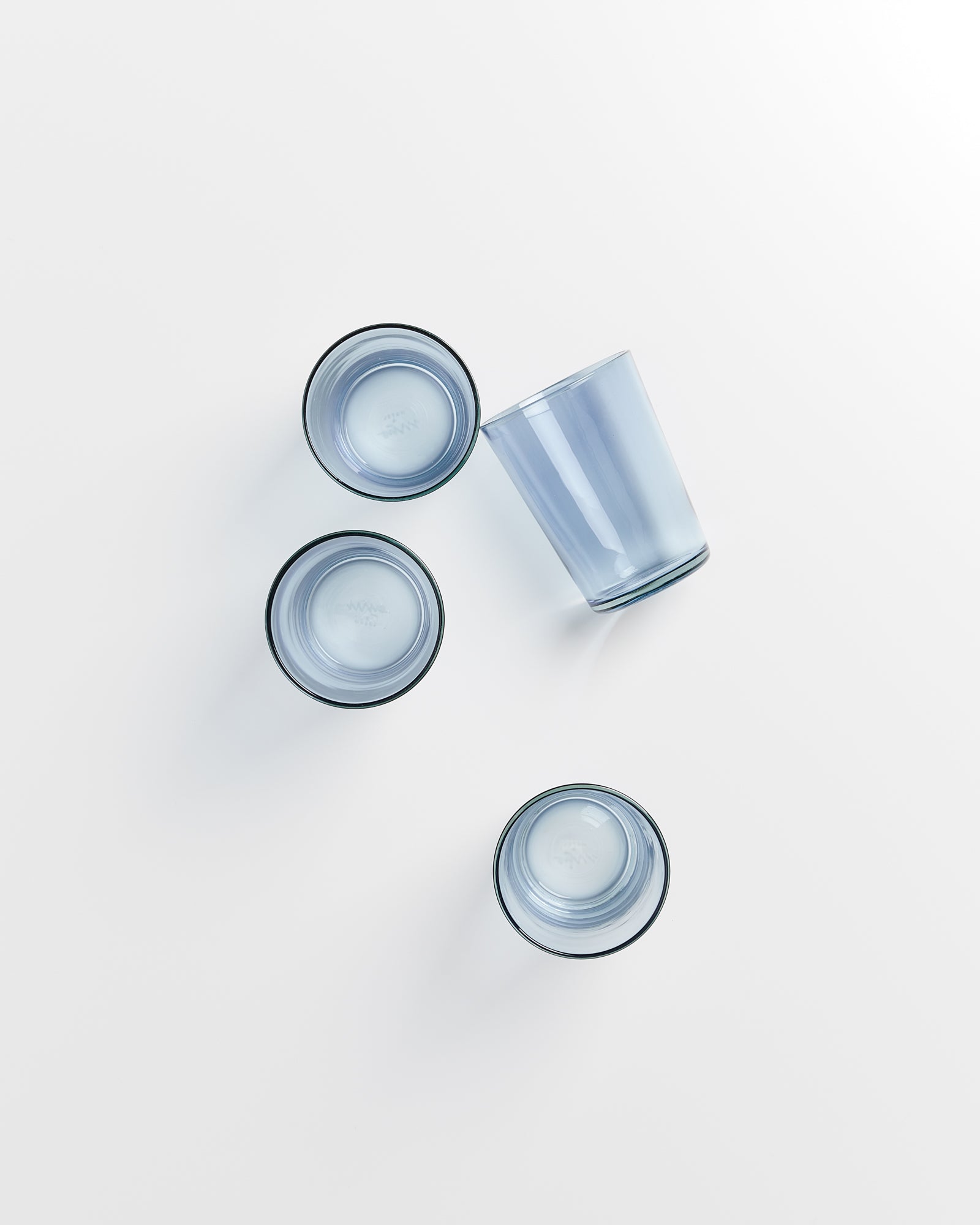 Vier blaue Gläser, drei stehend und eins umgekippt, auf weißem Hintergrund liegend.