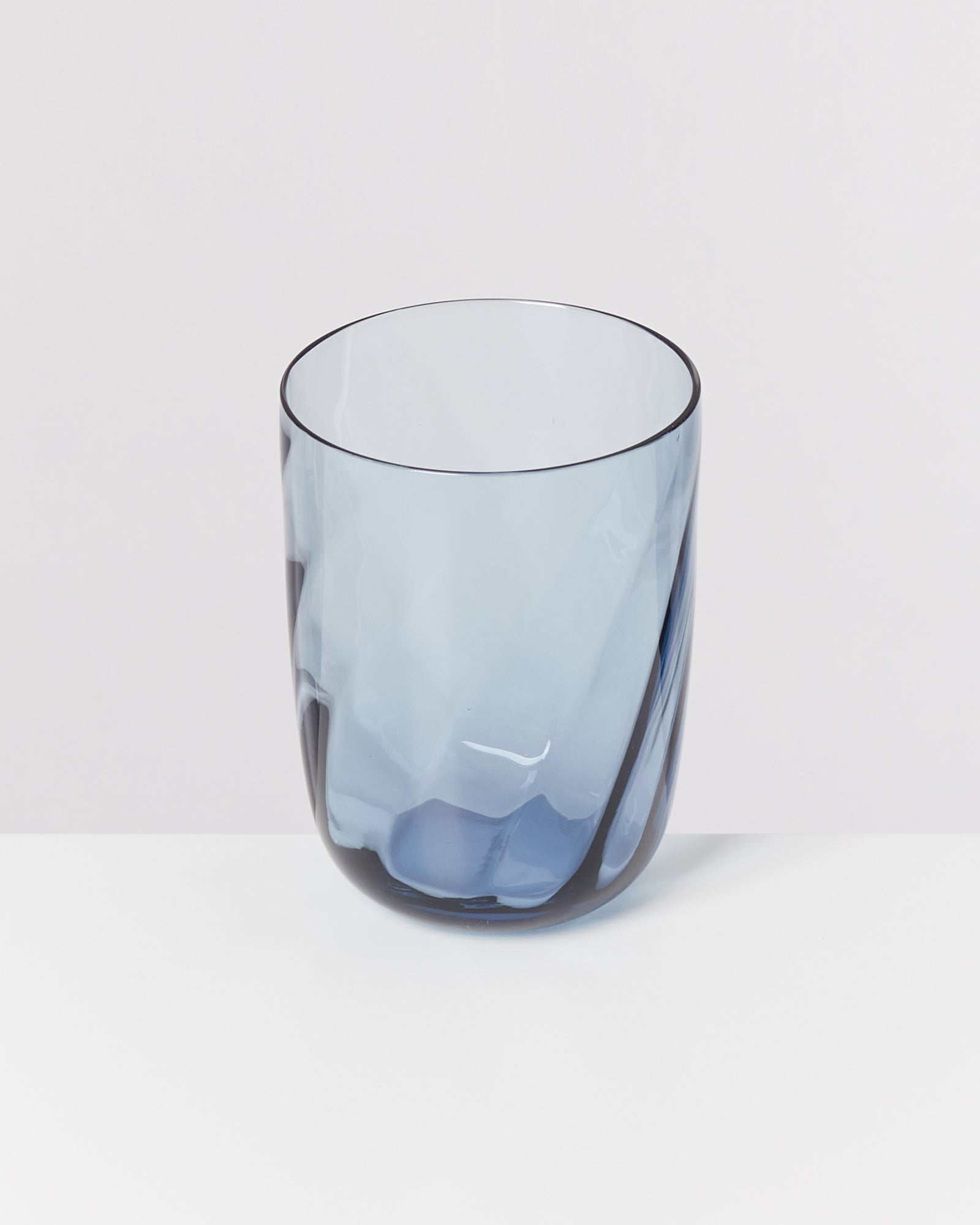Ein blaues, transparentes Glas mit geschwungener Oberfläche steht auf einem weißen Tisch vor einem hellen Hintergrund.