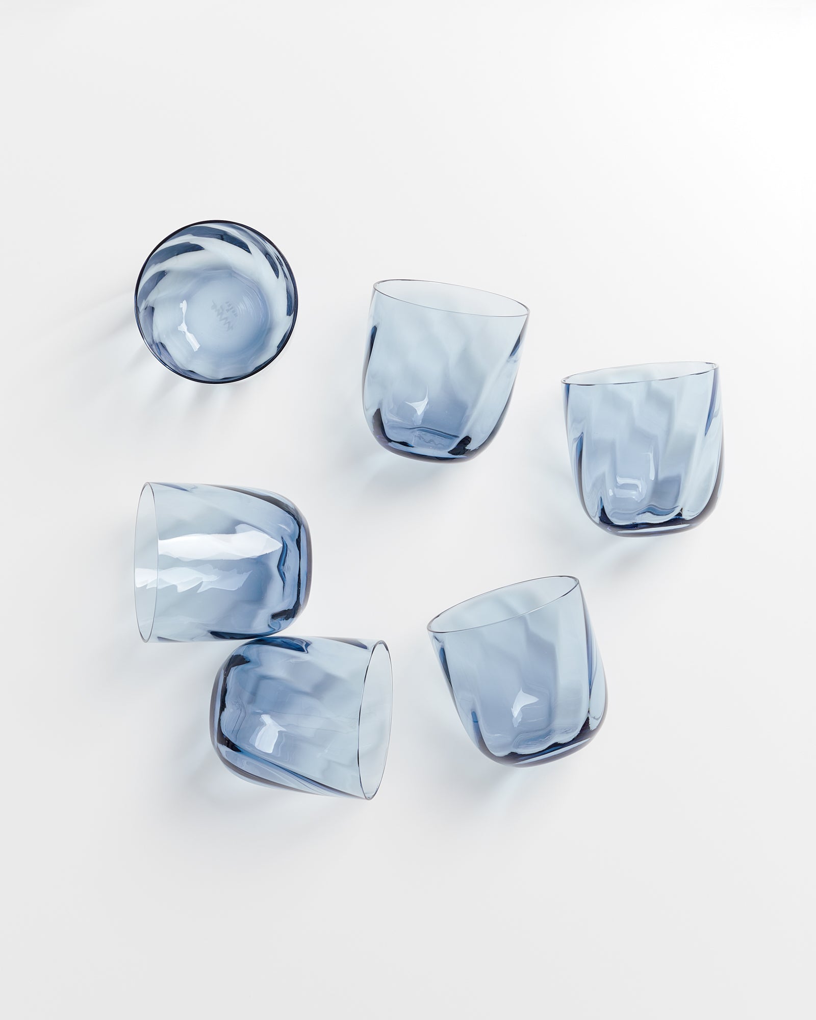 Sechs blaue, asymmetrische Trinkgläser liegen verstreut auf einer weißen Oberfläche.