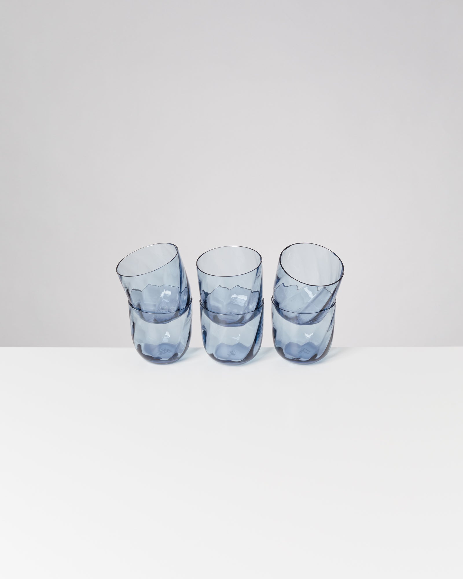Drei blaue, wellenförmige Trinkgläser stehen geneigt auf einer weißen Oberfläche vor einem grauen Hintergrund.