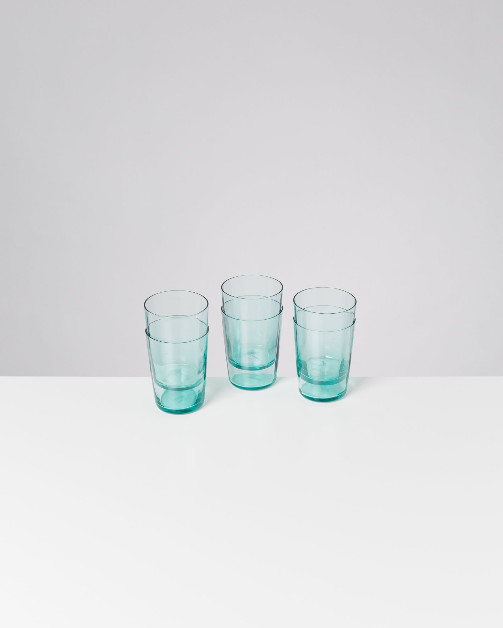 Drei leere, durchsichtige Glasbecher stehen auf einem weißen Tisch vor einem neutralen Hintergrund.