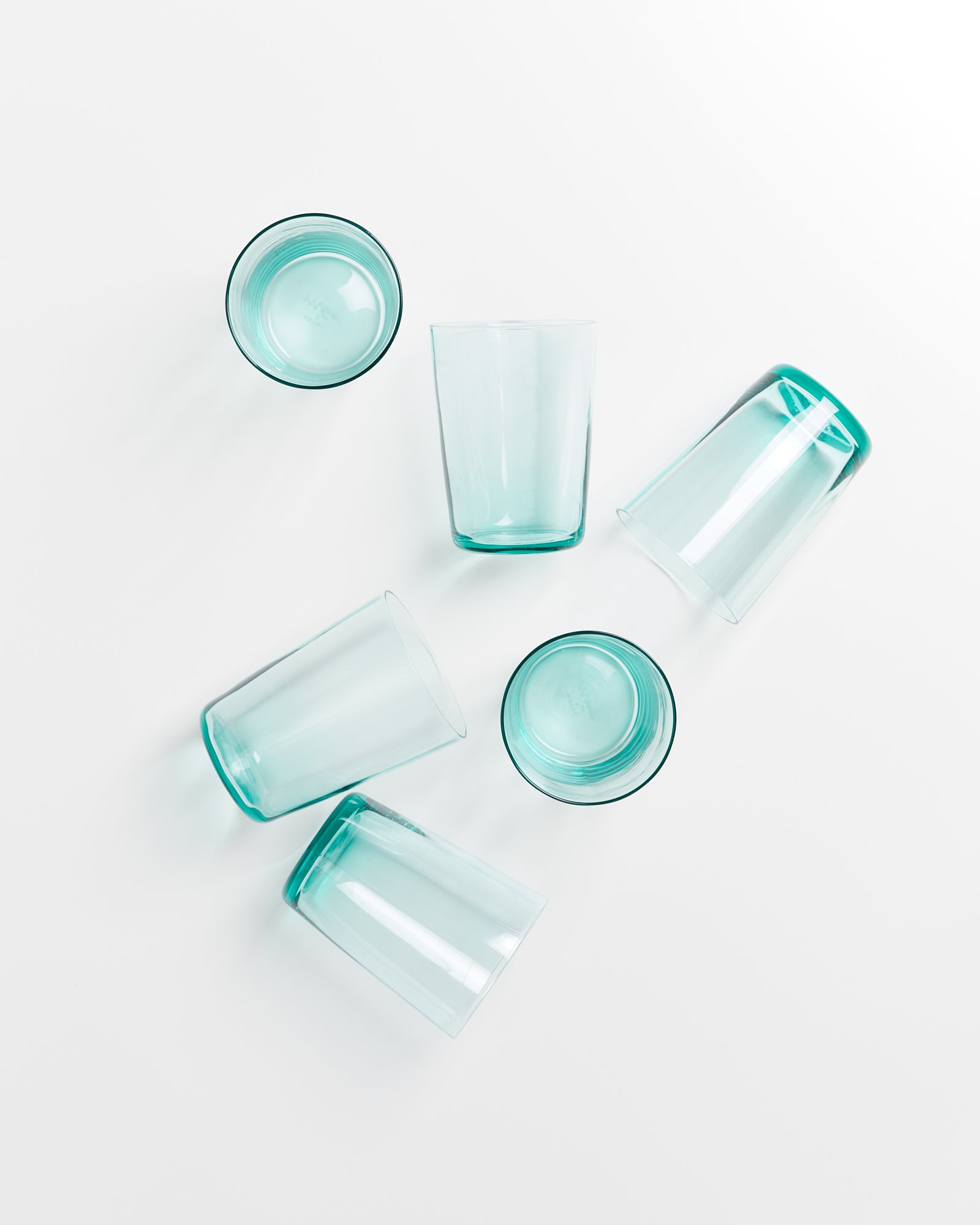 Fünf durchsichtige, türkisfarbene Trinkgläser liegen lose auf einer weißen Oberfläche.