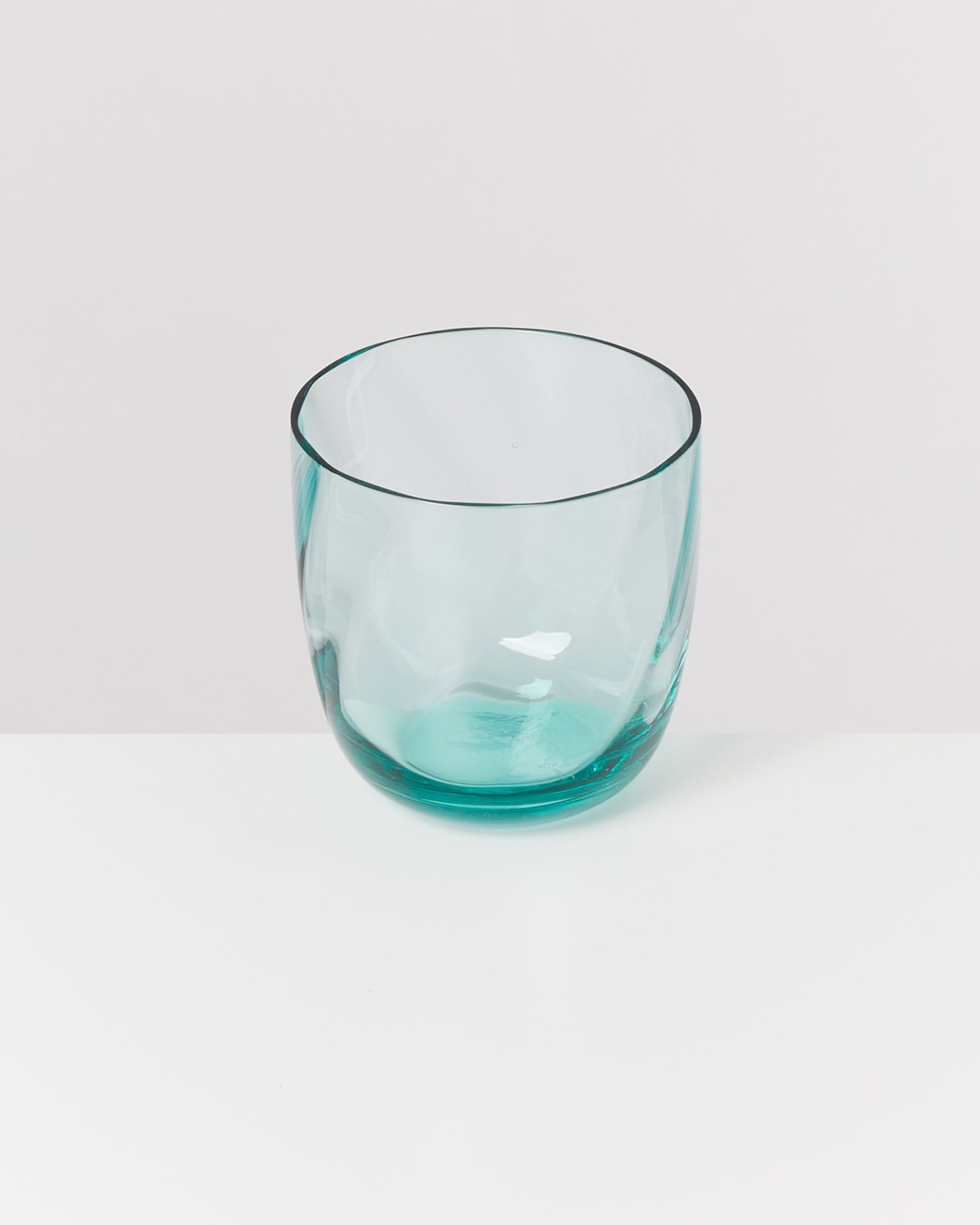 Ein türkisfarbenes Trinkglas steht auf einer weißen Oberfläche vor einem hellgrauen Hintergrund.