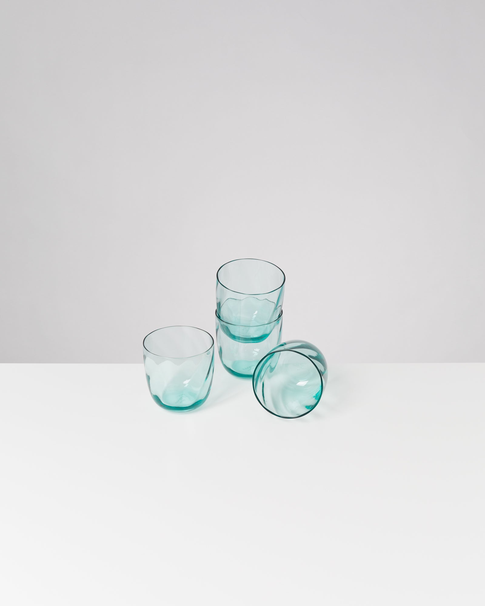 Drei grüne Gläser stehen und eines liegt auf der Seite auf einem weißen Tisch vor grauem Hintergrund.