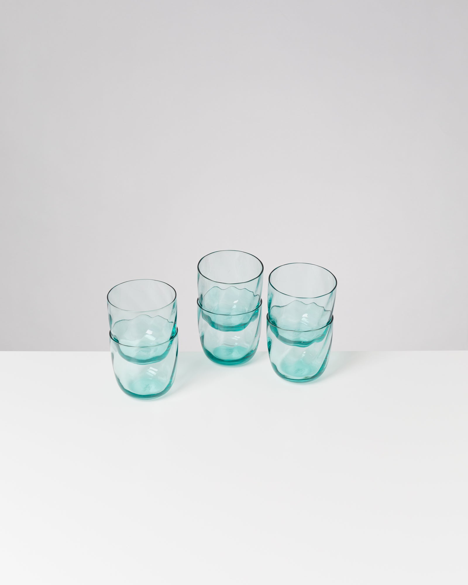 Drei türkisfarbene, ineinandergestapelte Trinkgläser auf einem weißen Tisch vor neutralem Hintergrund.