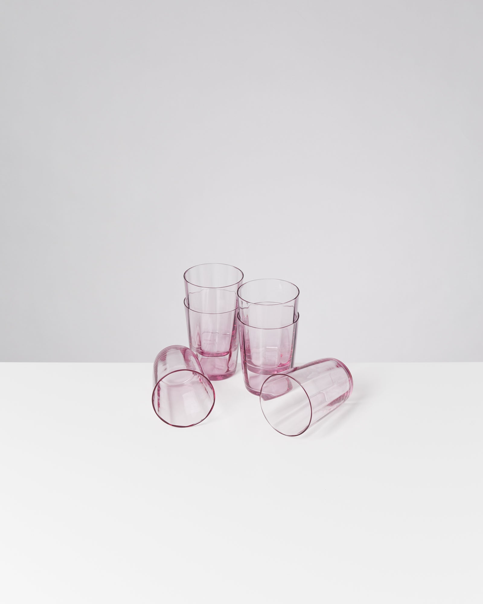 Fünf rosa Gläser stehen und liegen auf einem weißen Tisch vor grauem Hintergrund.