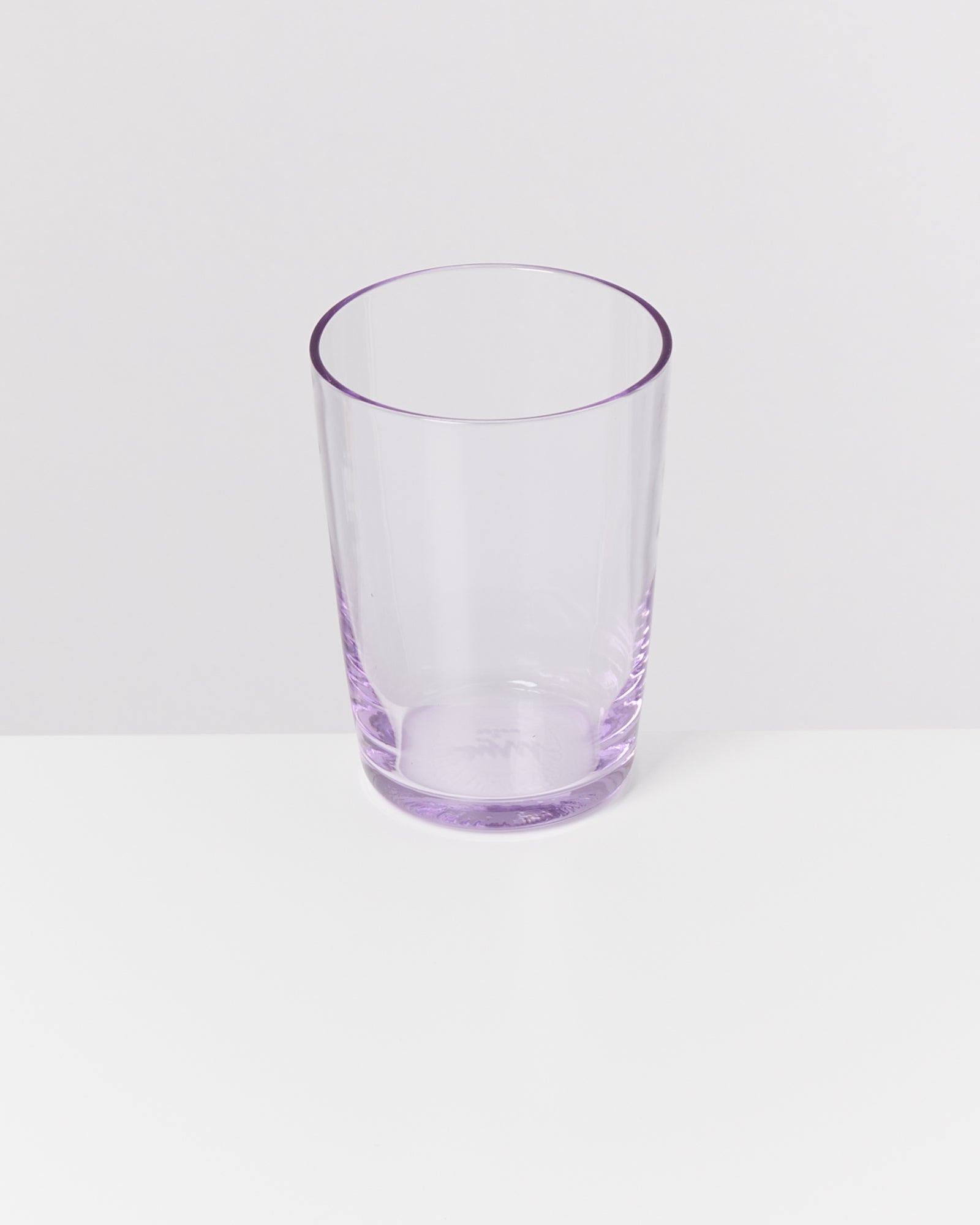 Ein transparentes, leicht lila gefärbtes Trinkglas steht auf einer weißen Oberfläche vor einem hellen Hintergrund.