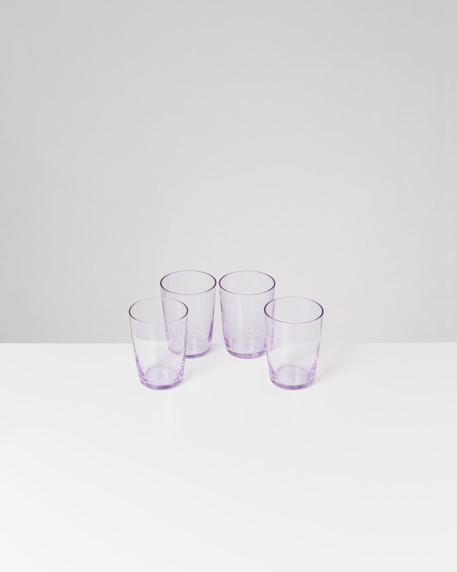 Vier durchsichtige, lilafarbene Gläser stehen auf einem weißen Tisch vor einem grauen Hintergrund.