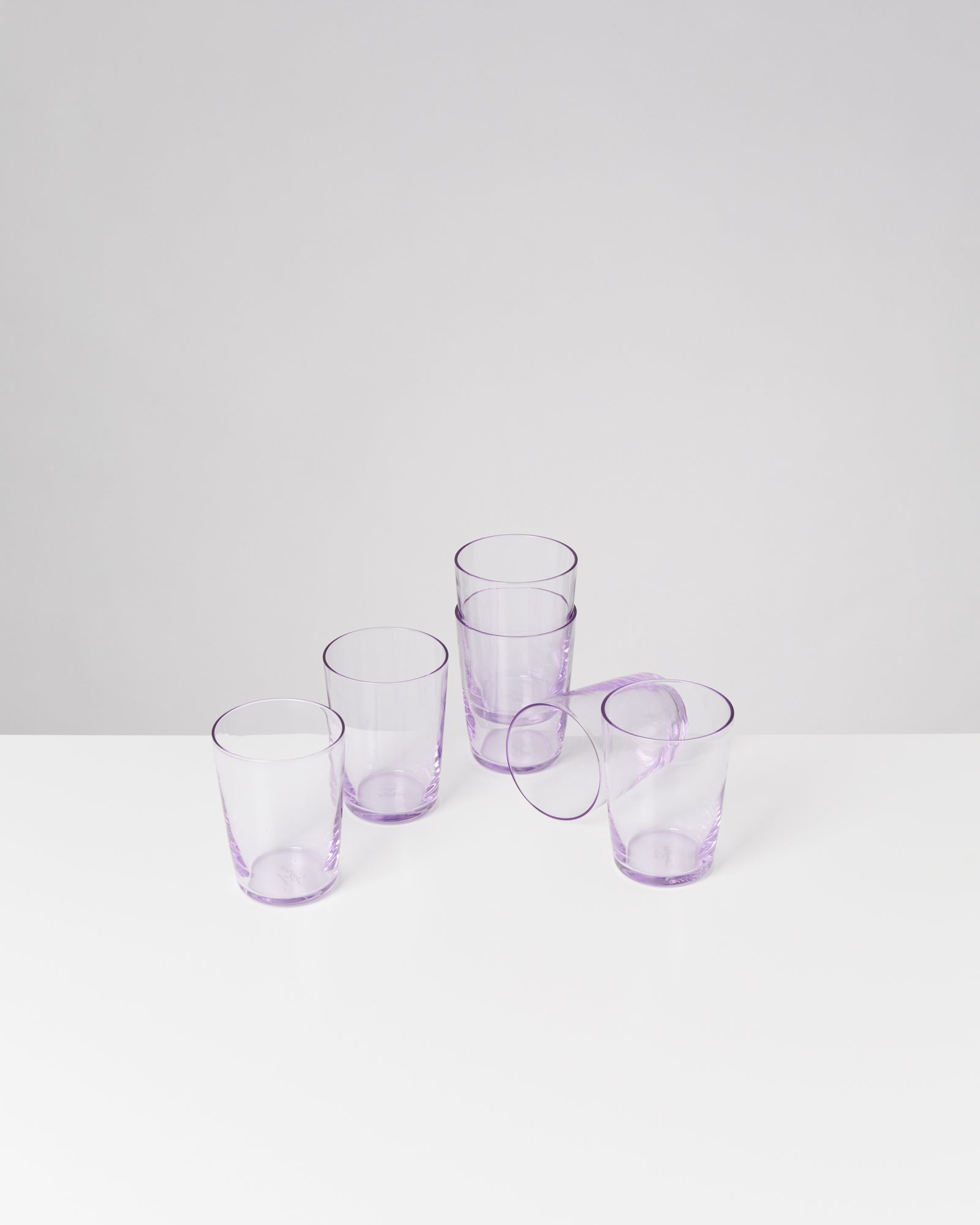 Fünf lila durchsichtige Gläser stehen und liegen auf einem weißen Tisch vor grauem Hintergrund.