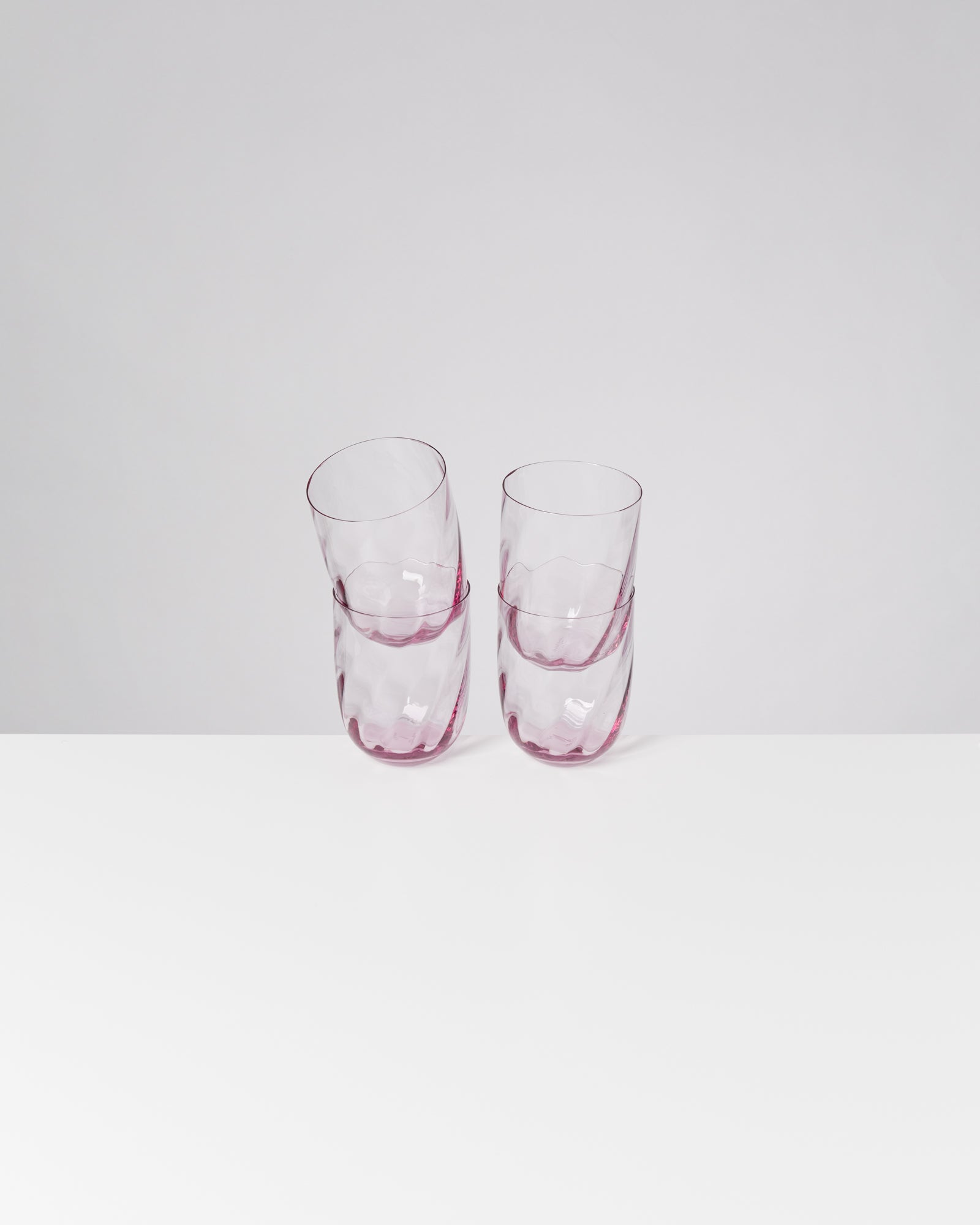 Vier gestapelte, blassrosa, facettierte Gläser stehen auf einer weißen Fläche vor einem grauen Hintergrund.