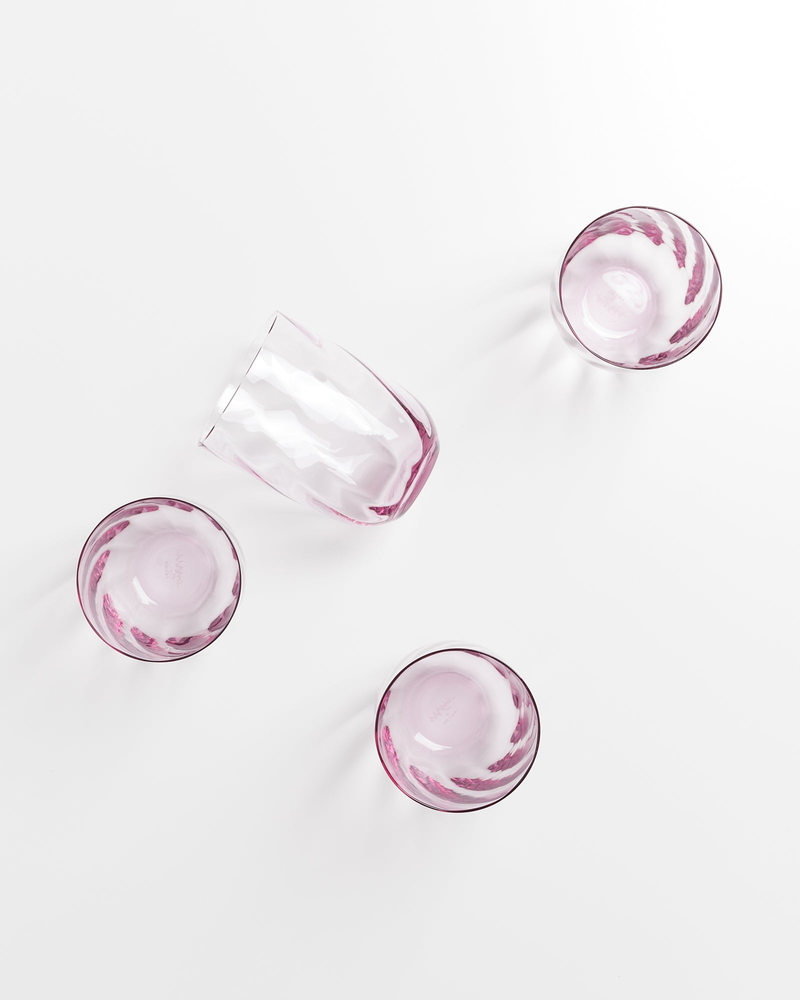 Vier rosafarbene Gläser, eines davon umgekippt, auf einer weißen Oberfläche.