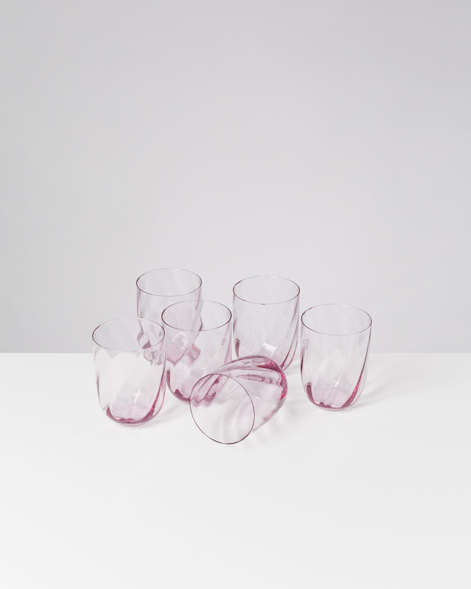 Sechs rosa Gläser, eines liegt auf der Seite, auf einer weißen Oberfläche vor einem hellen Hintergrund.