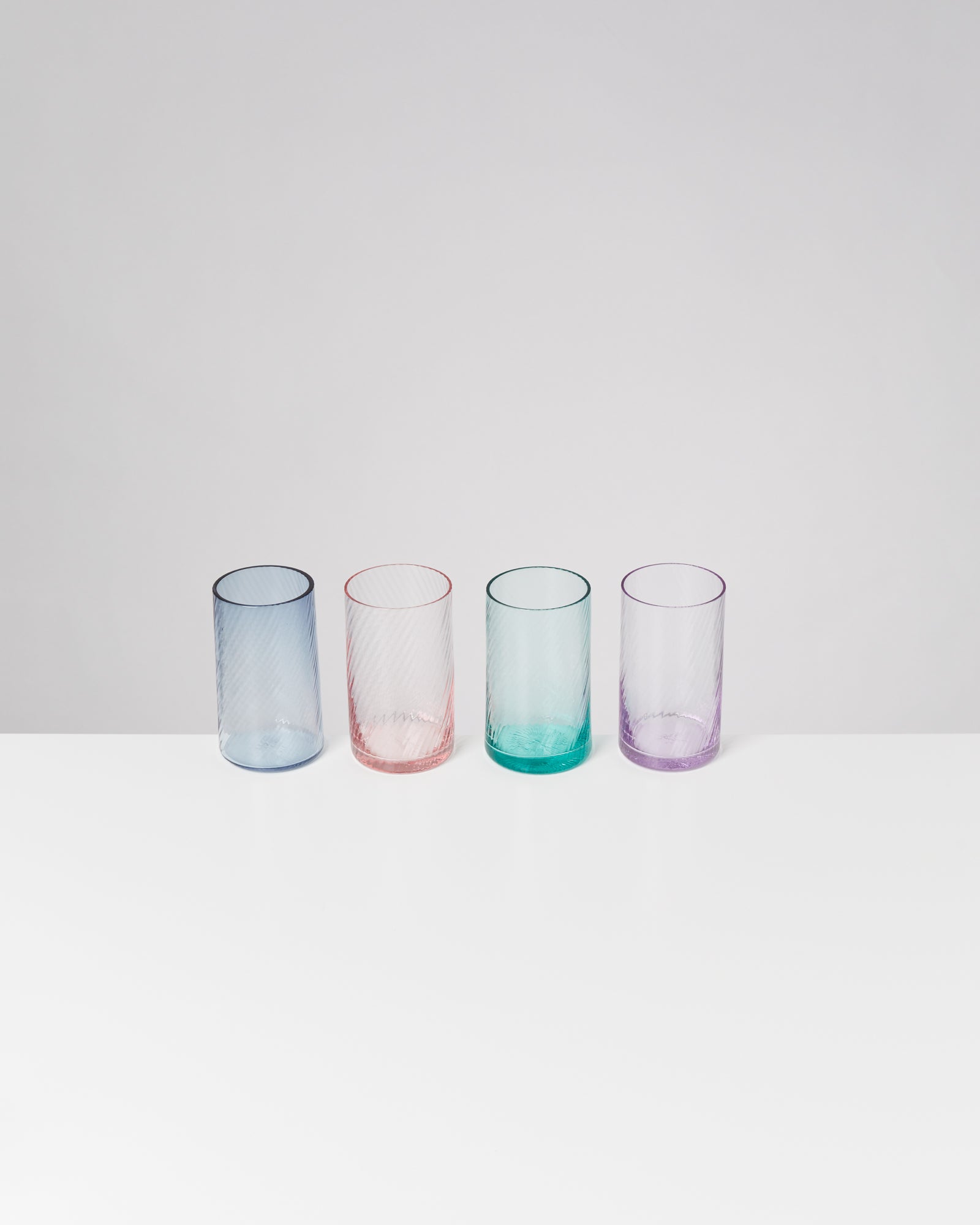 Vier bunte Gläser in Blau, Rosa, Grün und Violett stehen auf einer weißen Oberfläche vor einem grauen Hintergrund.