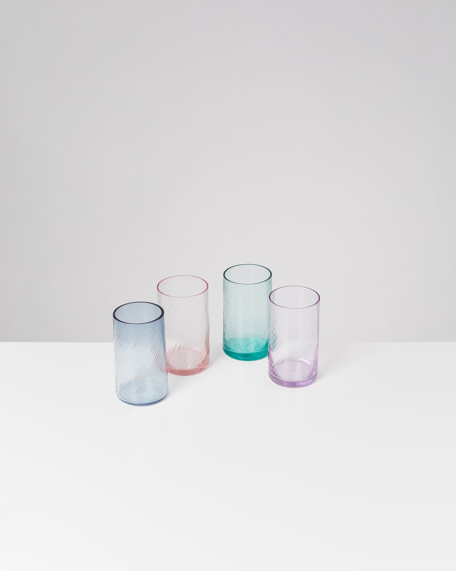 Vier bunte trinkgläser in Blau, Rosa, Grün und Lila auf einem weißen Tisch vor einem grauen Hintergrund.