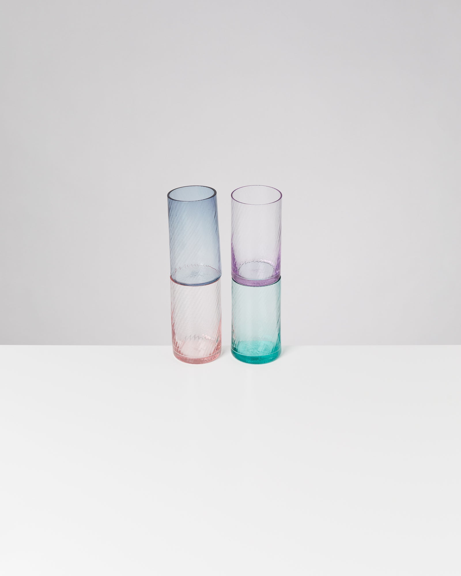 Vier bunte, durchsichtige Gläser stehen paarweise übereinander auf einem weißen Tisch vor einem grauen Hintergrund.