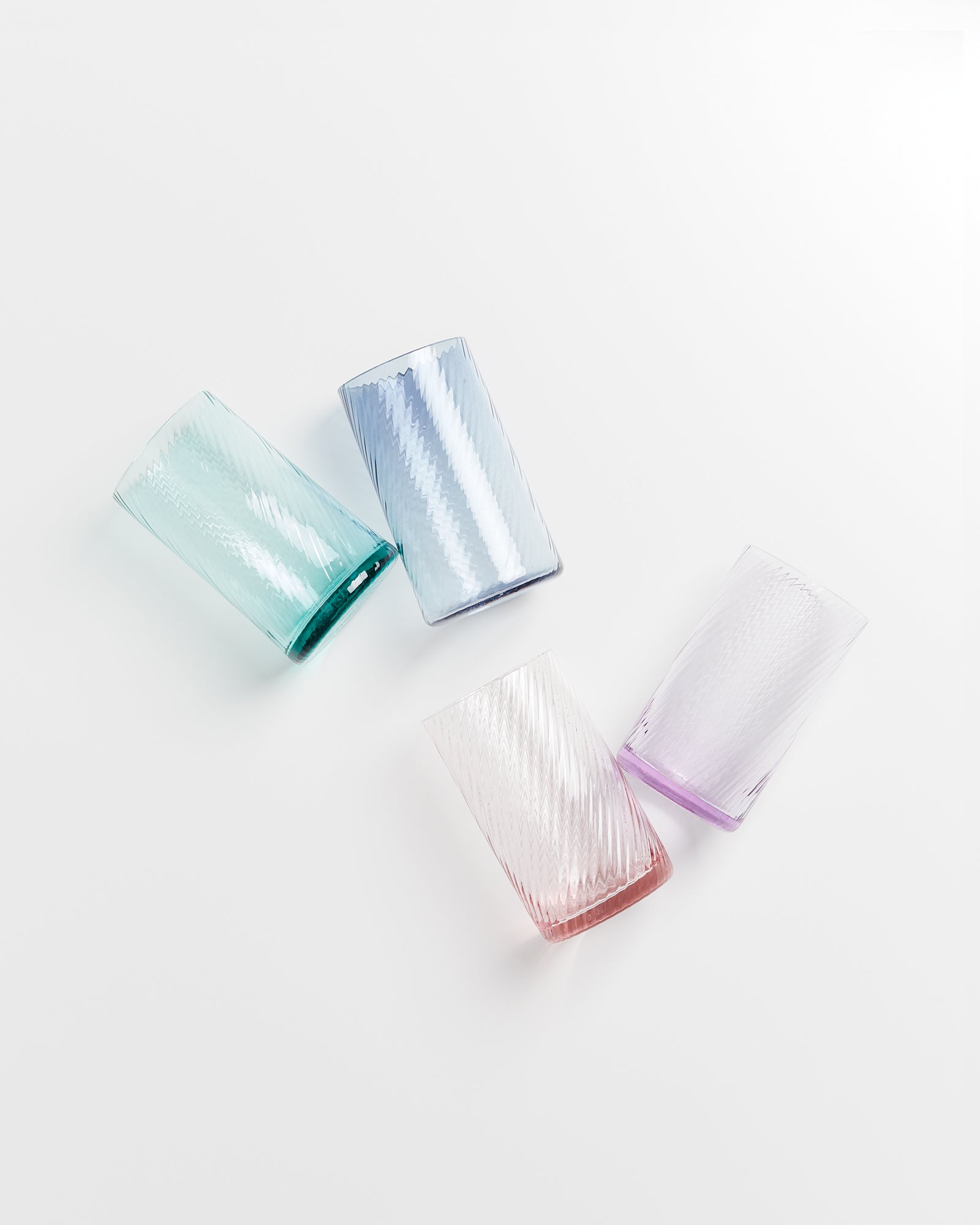 Vier bunte, strukturierte Trinkgläser liegen schräg auf einer weißen Oberfläche.