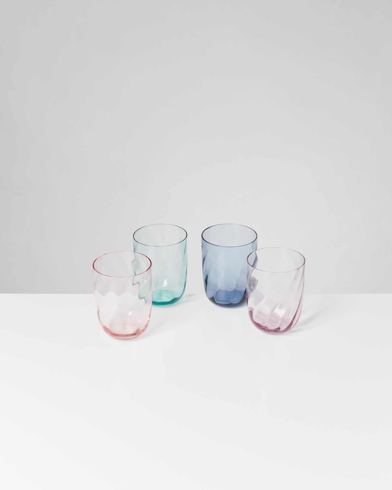 Vier bunte, gewellte Gläser in Pastellfarben stehen auf einer weißen Oberfläche vor einem neutralen Hintergrund.