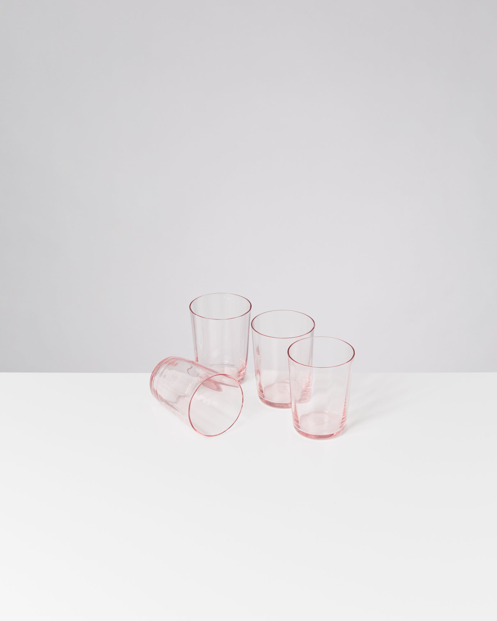 Vier transparente rosa Gläser, drei stehen aufrecht, eines liegt umgestürzt auf einer weißen Oberfläche.