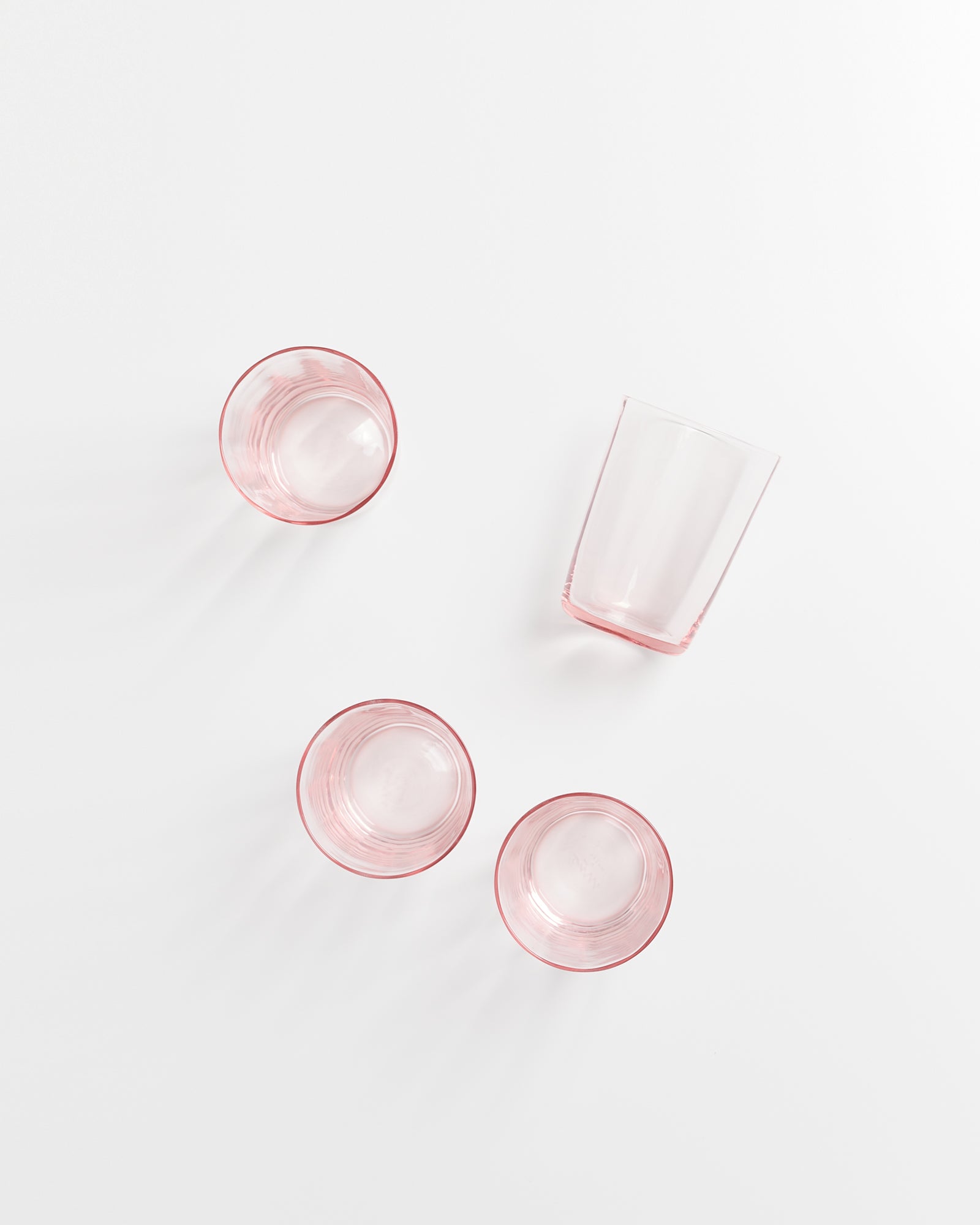 Vier rosafarbene Trinkgläser auf weißem Hintergrund, eins leicht gekippt.