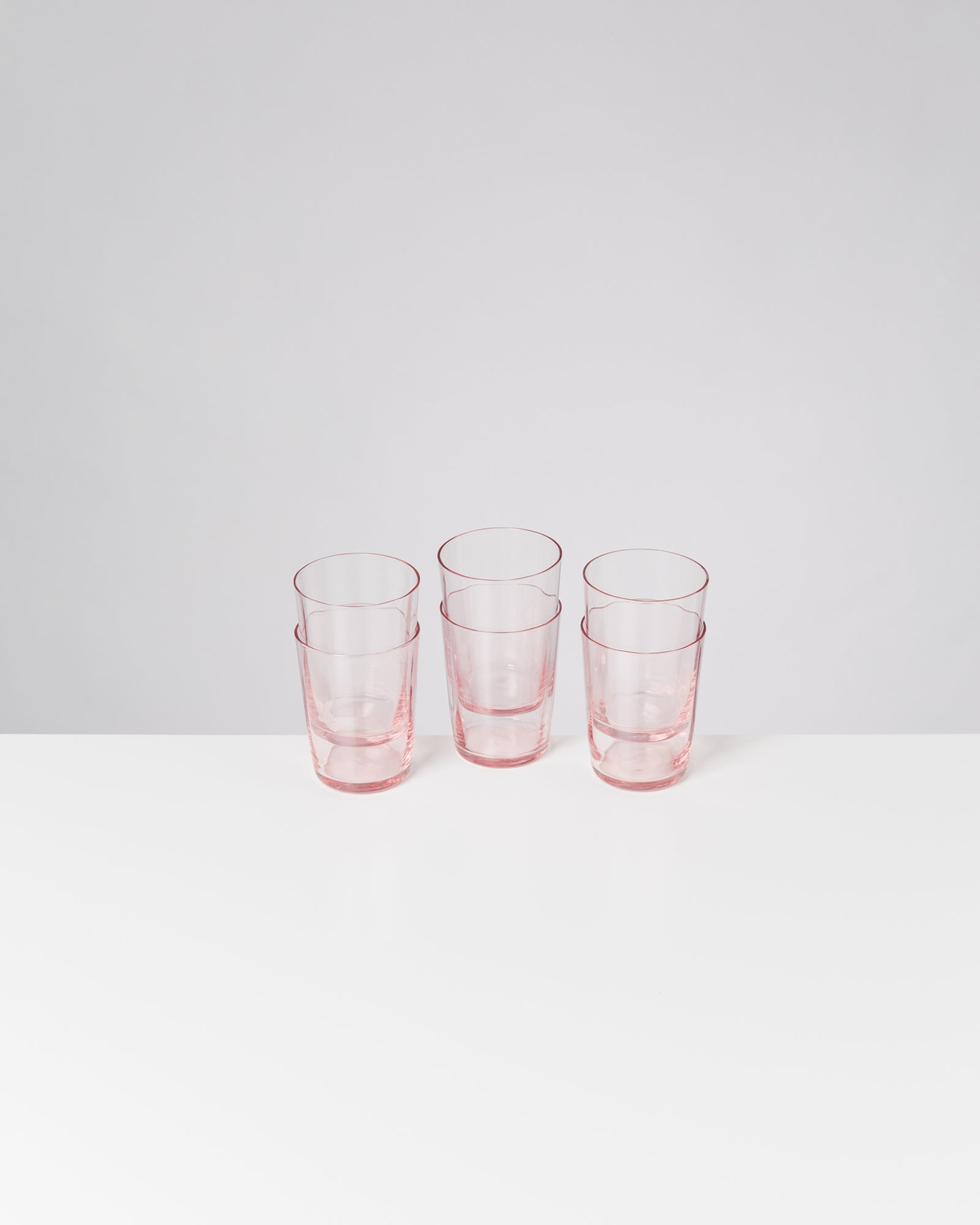 Fünf rosa Gläser stehen in zwei Reihen auf einem weißen Tisch vor einem grauen Hintergrund.