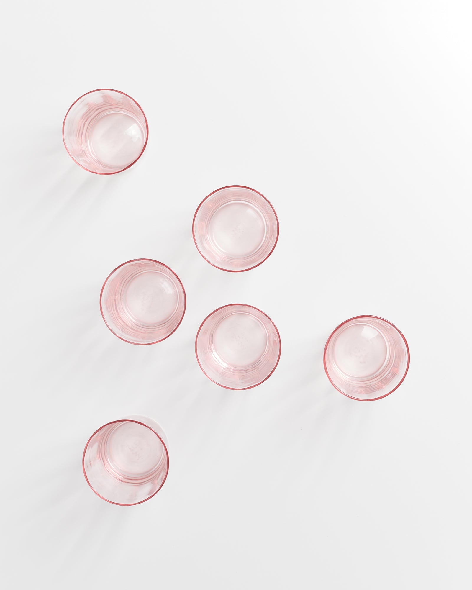 Sechs leere, rosafarbene Gläser stehen auf einer weißen Fläche.