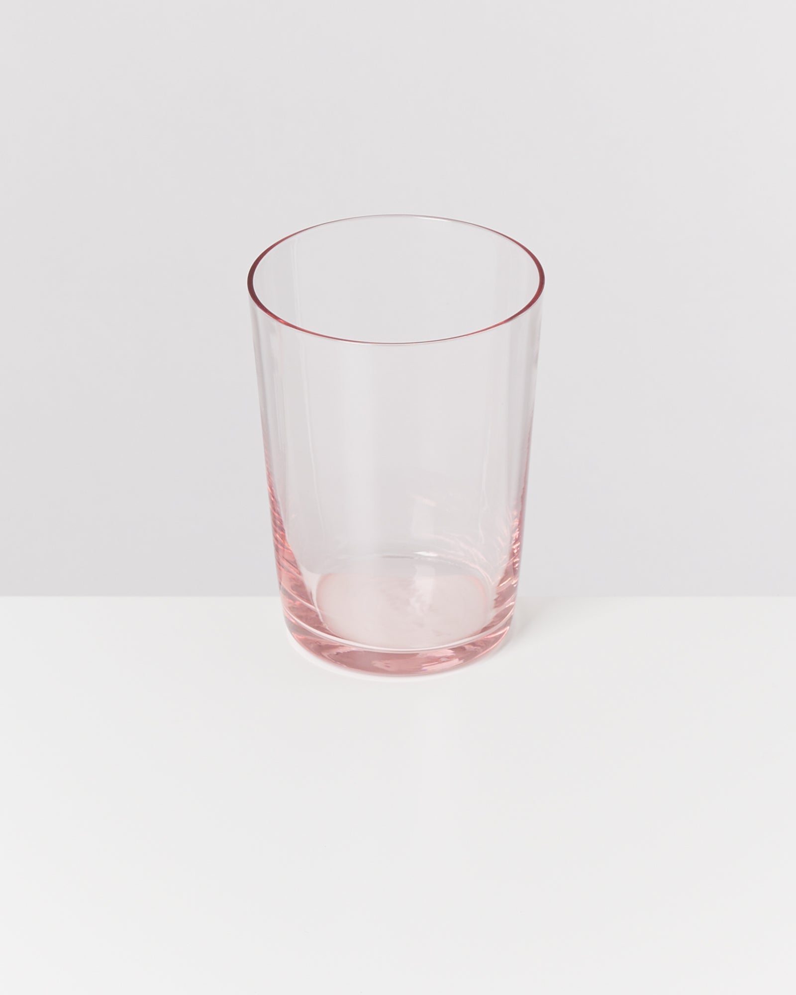 Ein durchsichtiges Glas mit rosa Schimmer steht auf einem weißen Tisch vor einem grauen Hintergrund.