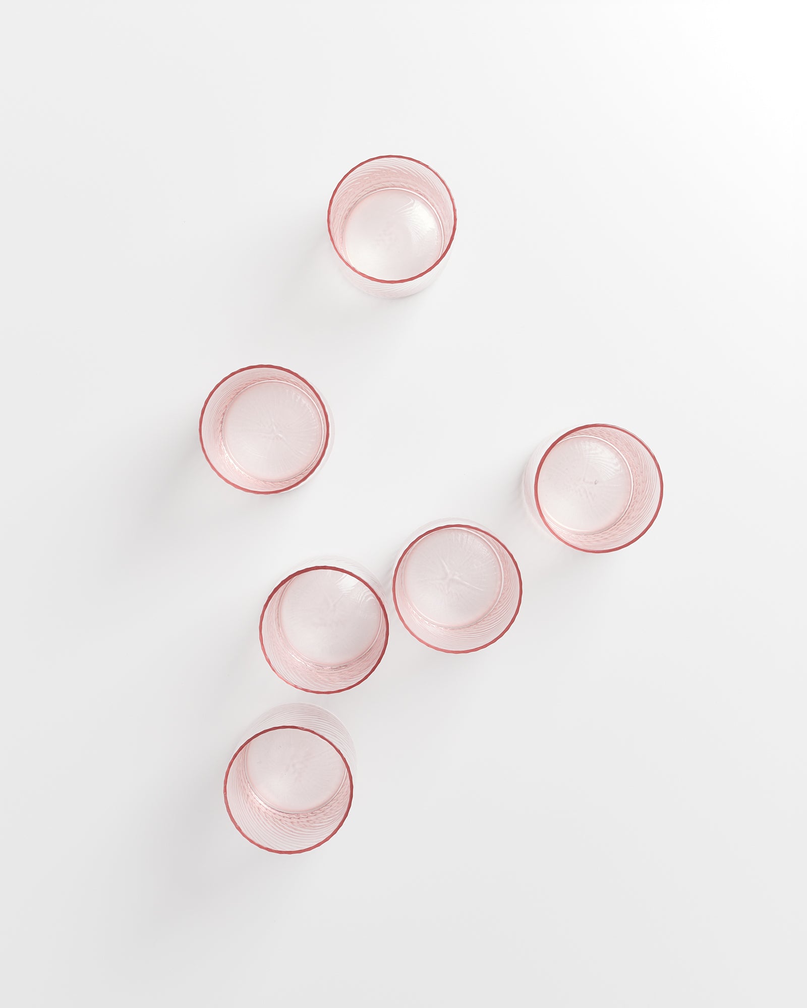 Sechs rosafarbene Gläser stehen in einer diagonalen Linie auf einer weißen Oberfläche.