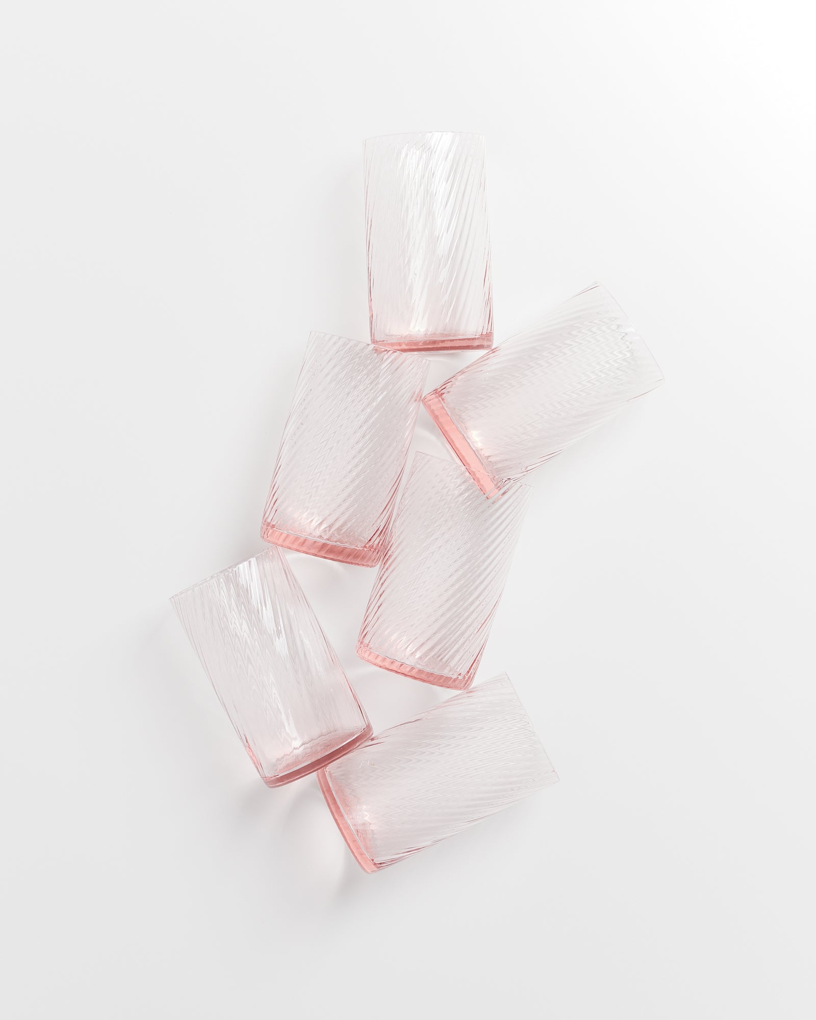 Sechs leicht rosa gefärbte, geriffelte Gläser liegen schräg nebeneinander auf einer weißen Oberfläche.